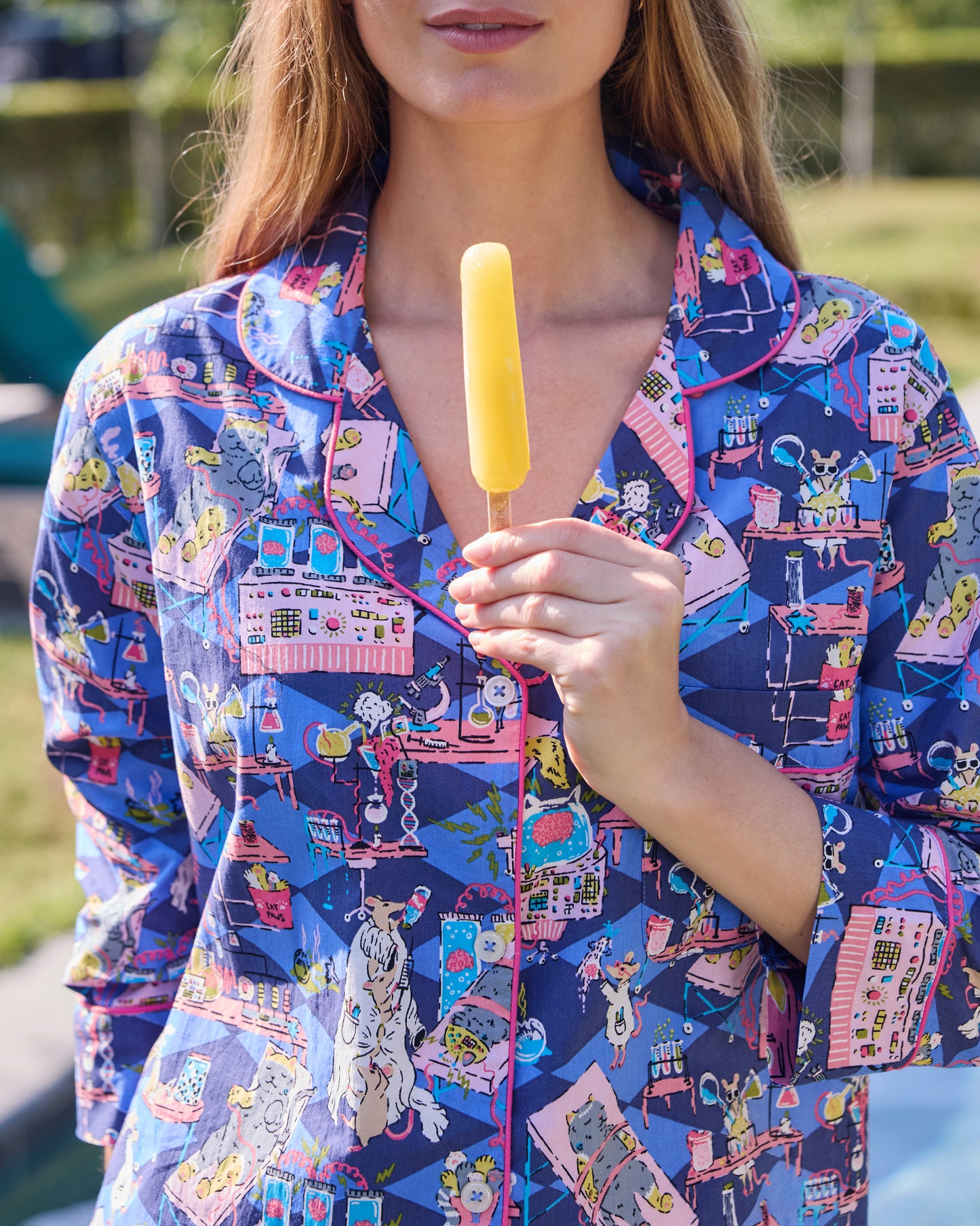 Close view of a blue button-down sleep shirt with a colorful mice-and-laboratory print, while a person holds a yellow popsicle.