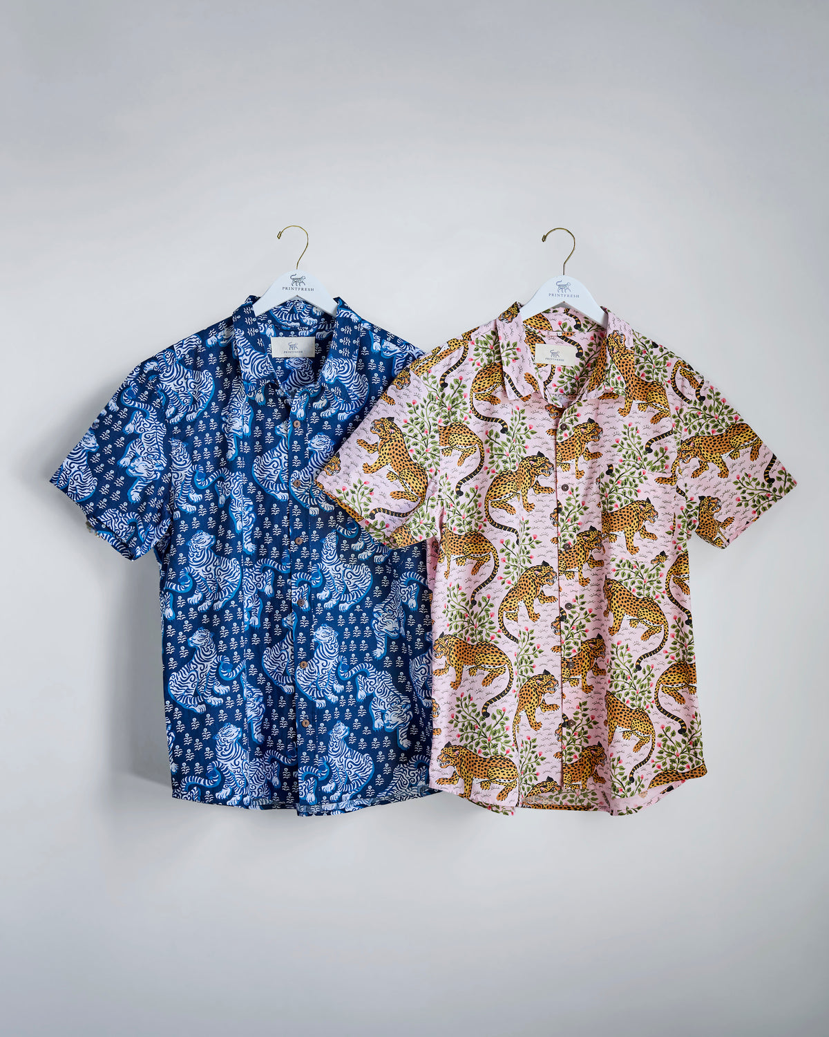 Two short-sleeve button-down shirts on hangers: one blue with a white tiger pattern and one pink with a leopard pattern and green foliage.
