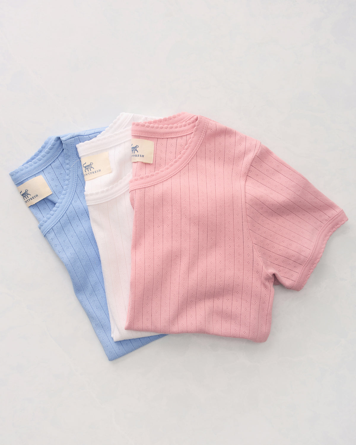 Three short-sleeve pointelle tees in blue, white, and pink, arranged overlapping on a light background.