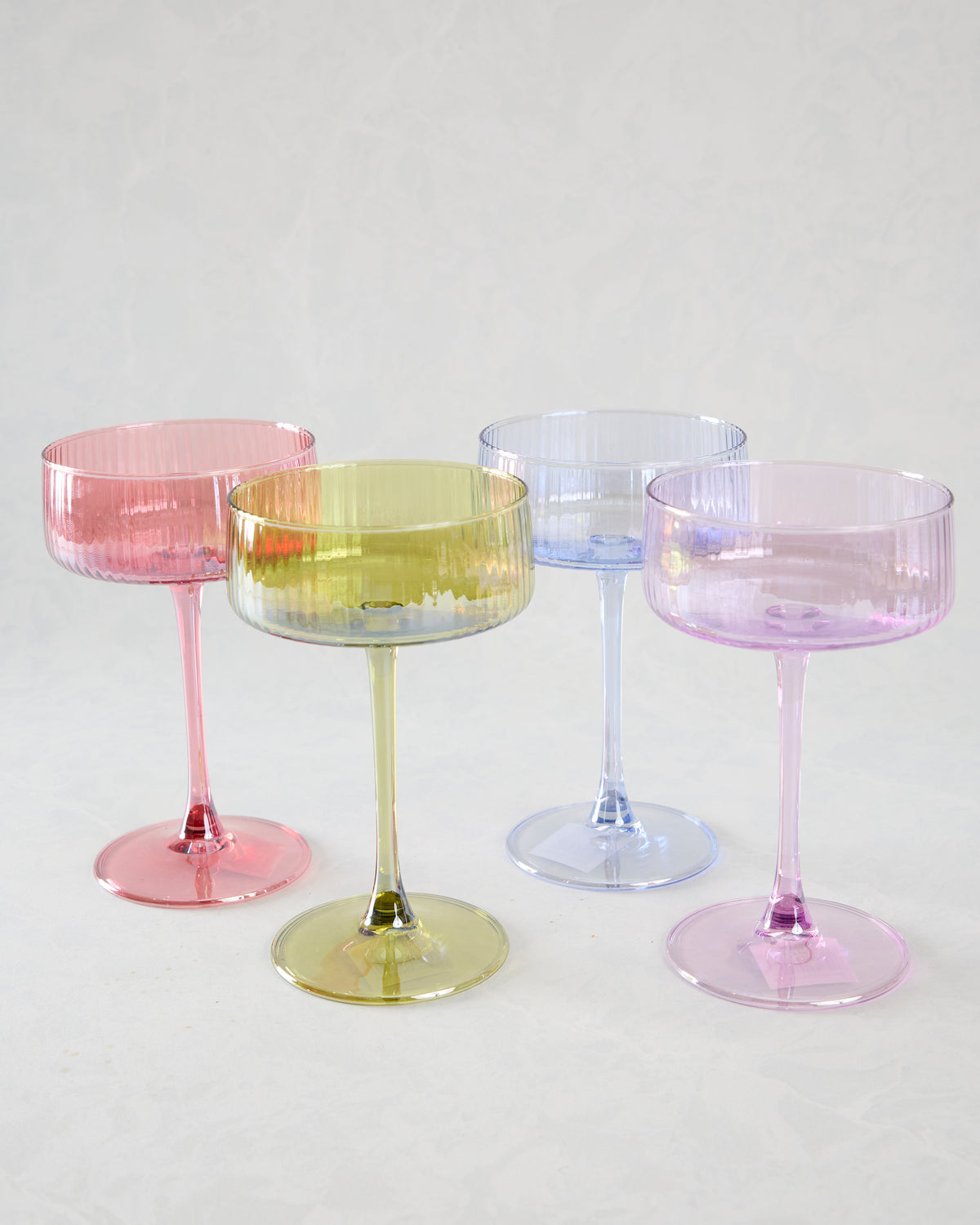 Four assorted translucent cocktail coupes with fluted bowls and slender stems, shown in pink, yellow, blue, and purple.