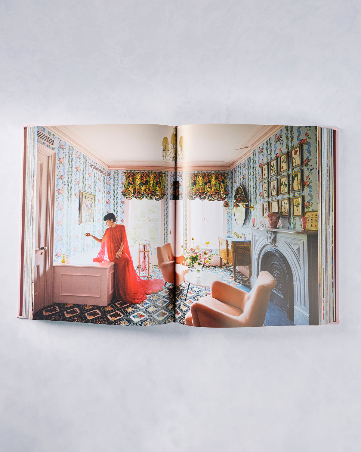 Open Architectural Digest book showing a colorful living room with floral wallpaper, framed art, a fireplace, and a person in a red dress.
