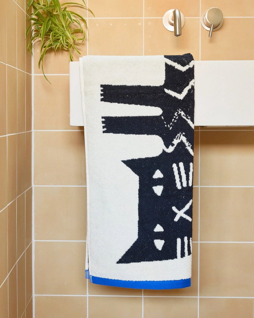 White hand towel with a black cat illustration and blue edging hanging on a bathroom rail.