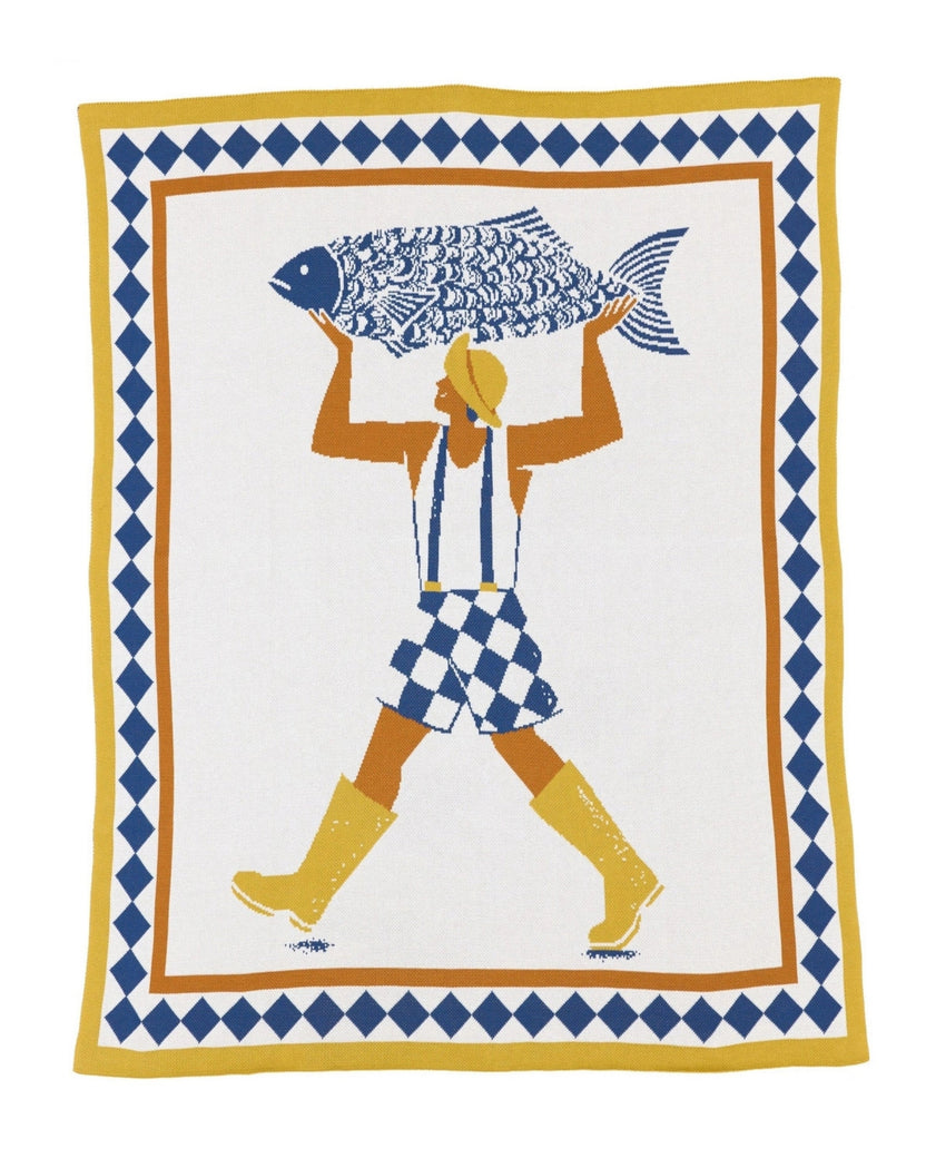 Un Beau Poisson knit throw showing a figure carrying a large fish, framed by blue diamond and yellow borders.