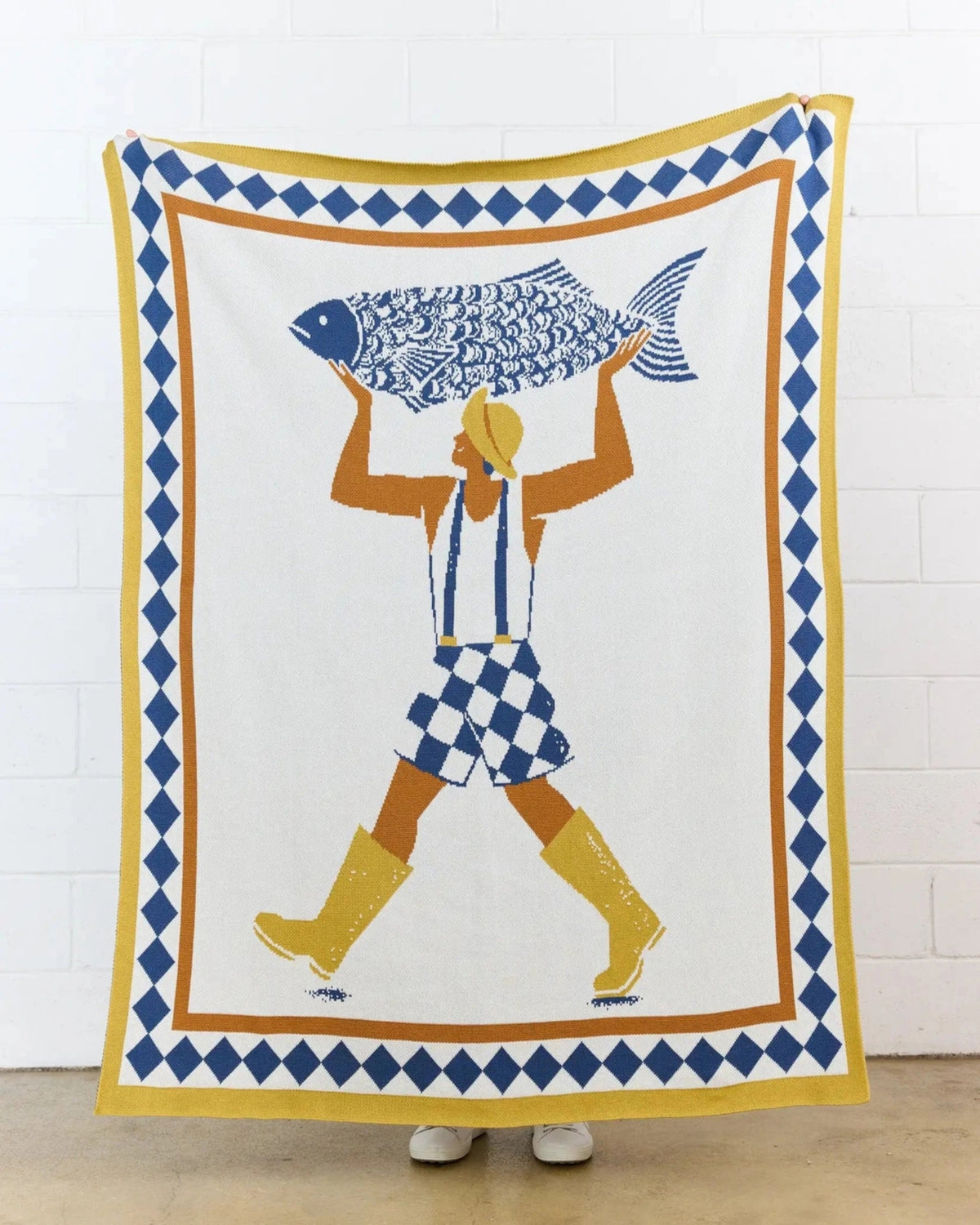 Un Beau Poisson knit throw hanging against a white brick wall, with a figure carrying a large fish.