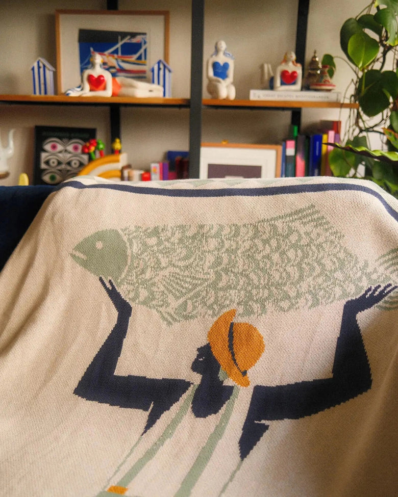 Close-up of the Un Beau Poisson throw draped over furniture, showing the figure carrying a fish.