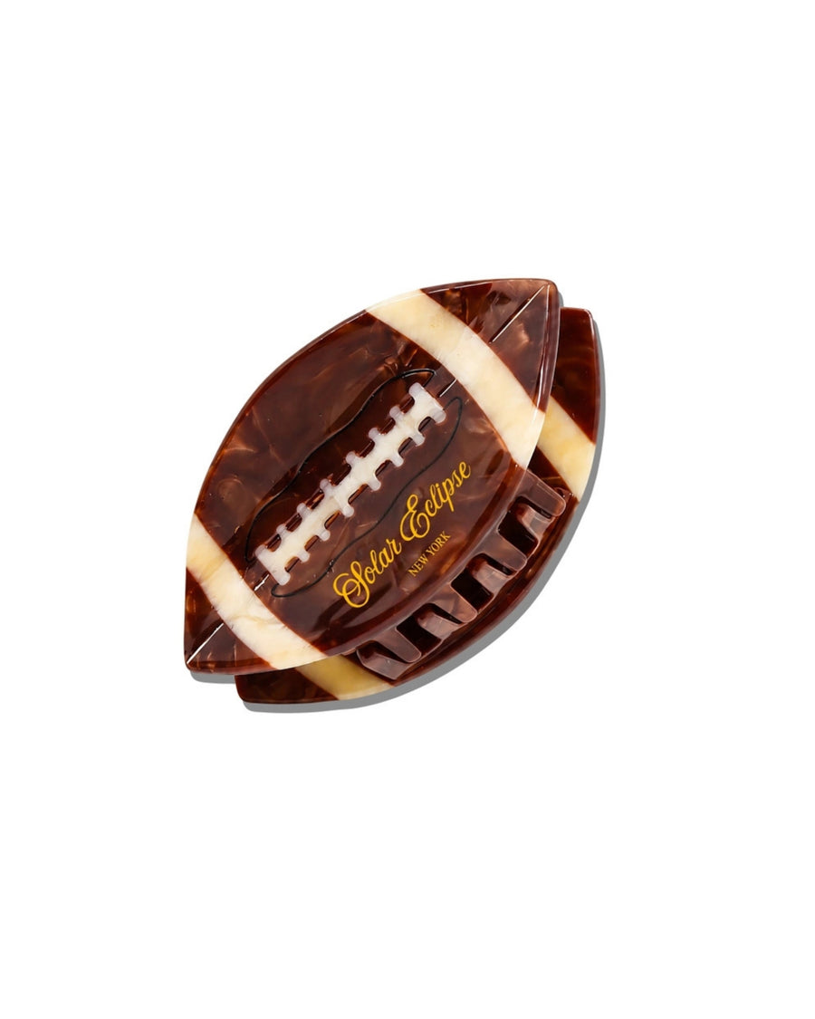 Angled view of a brown football-shaped claw hair clip showing its teeth, white stripes, and raised laces.