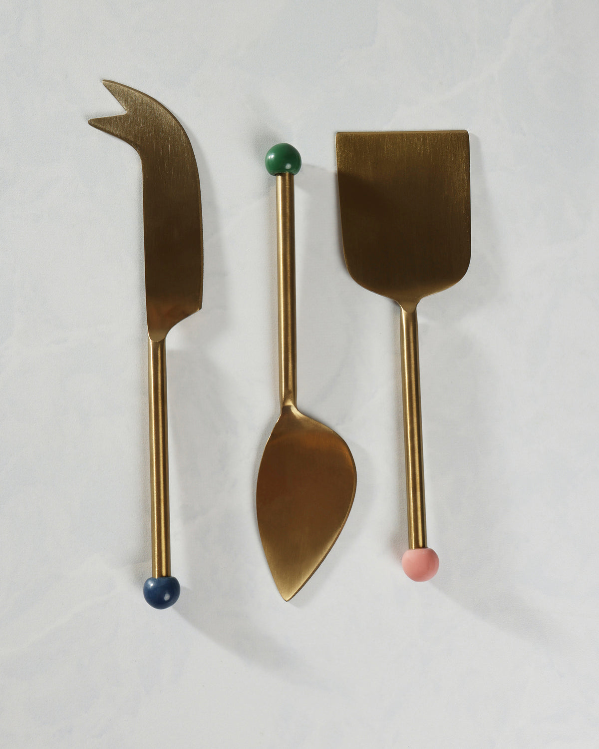Three-piece gold cheese knife set arranged vertically, including a pointed knife, a spoon-shaped utensil, and a wide flat server.