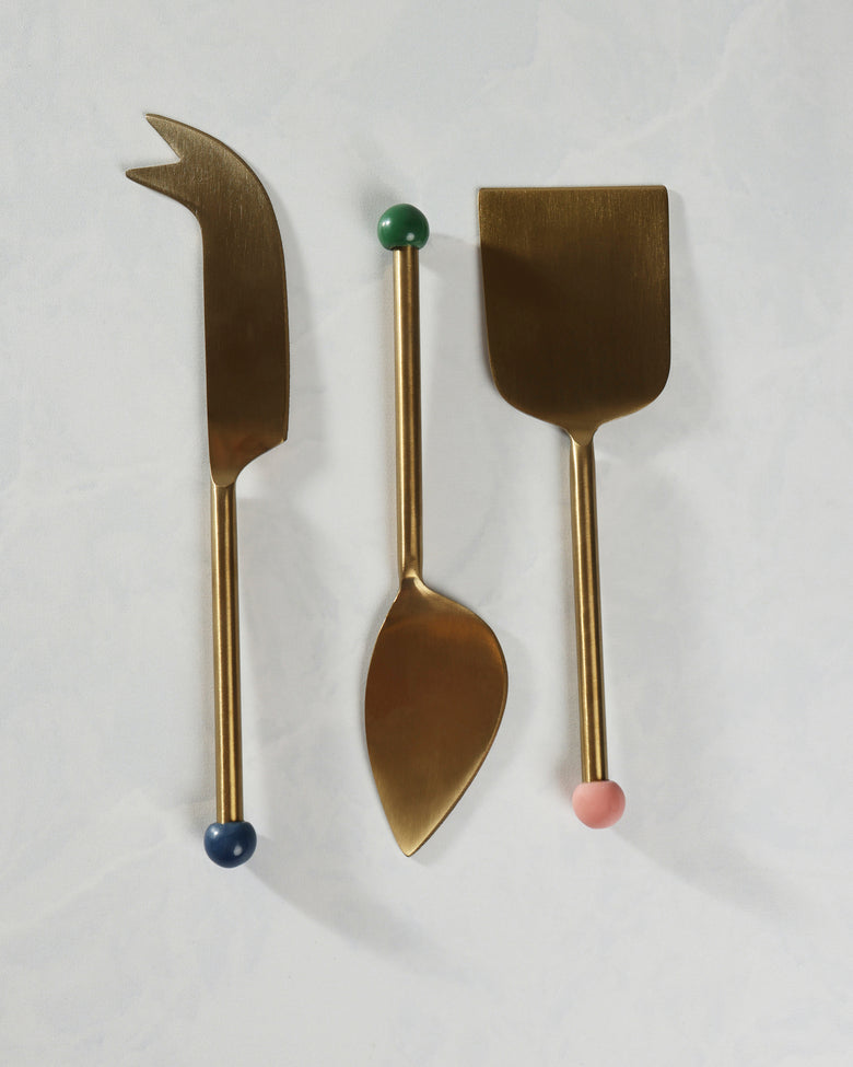 Three-piece gold cheese knife set arranged vertically, including a pointed knife, a spoon-shaped utensil, and a wide flat server.