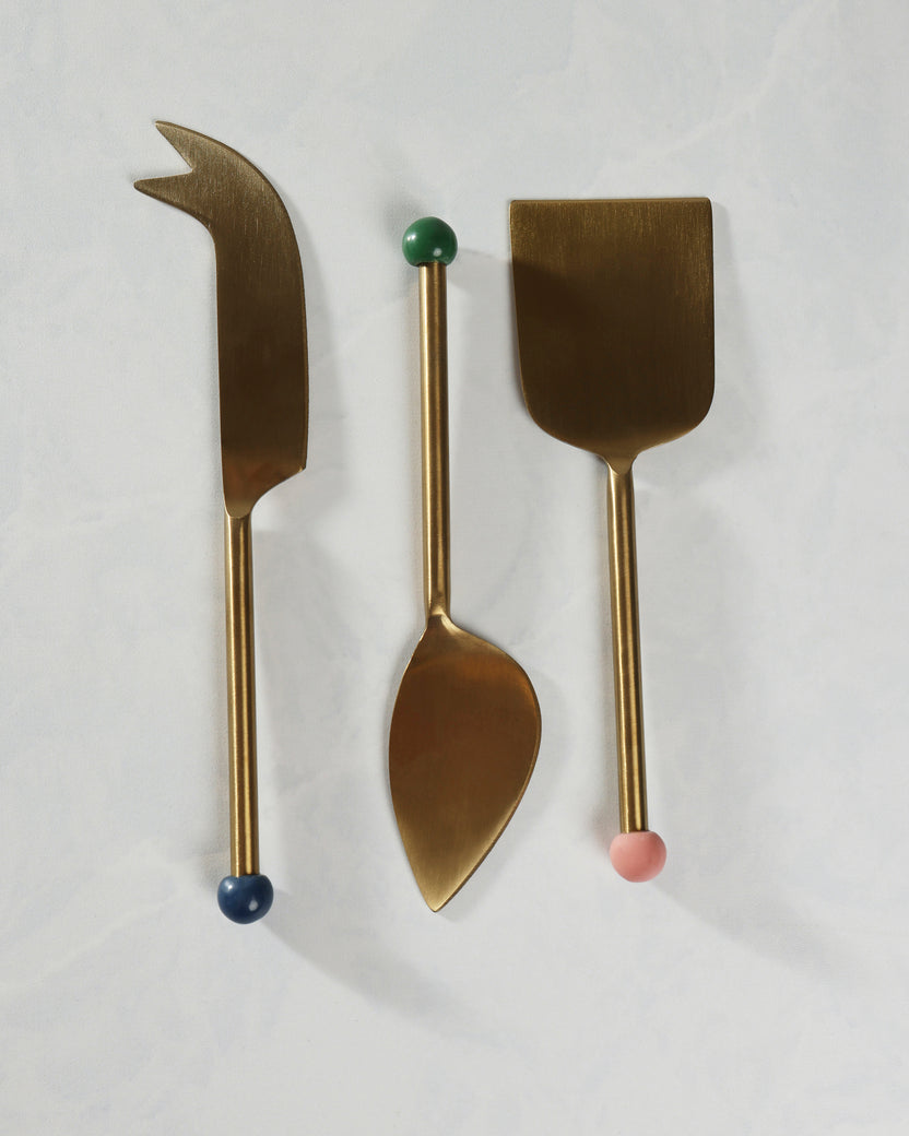 Three-piece gold cheese knife set arranged vertically, including a pointed knife, a spoon-shaped utensil, and a wide flat server.
