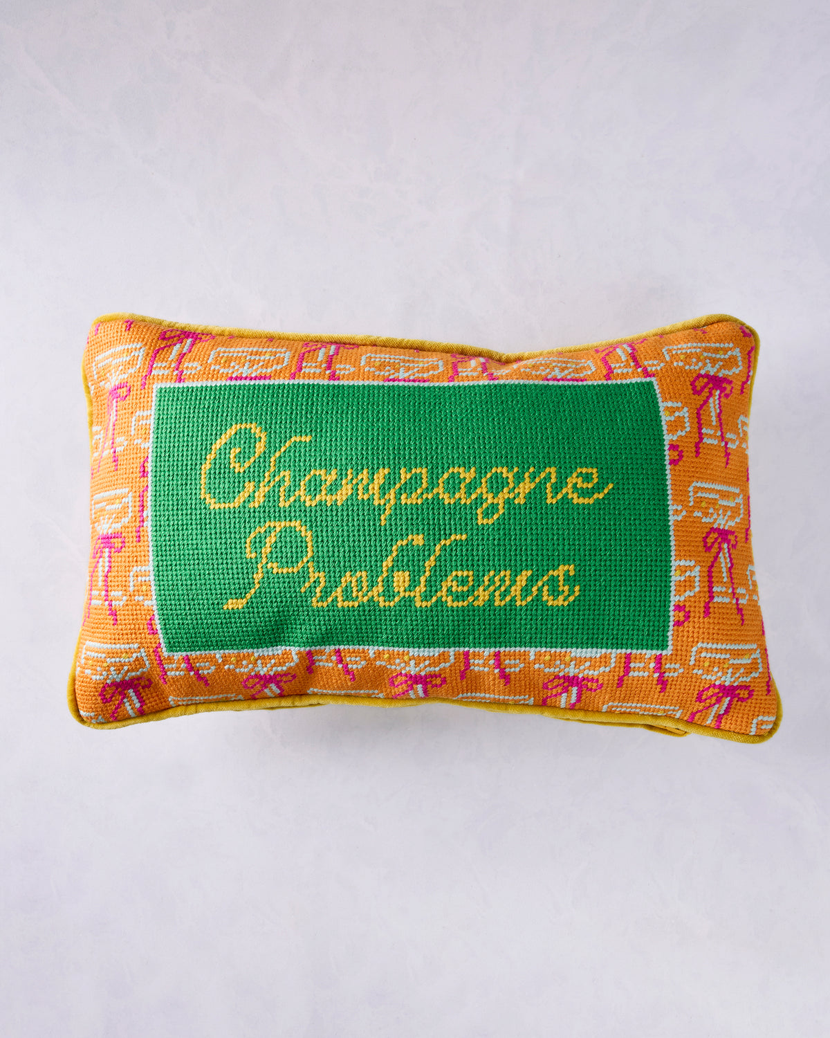 Needlepoint pillow with an orange patterned border and green center panel reading “Champagne Problems.”