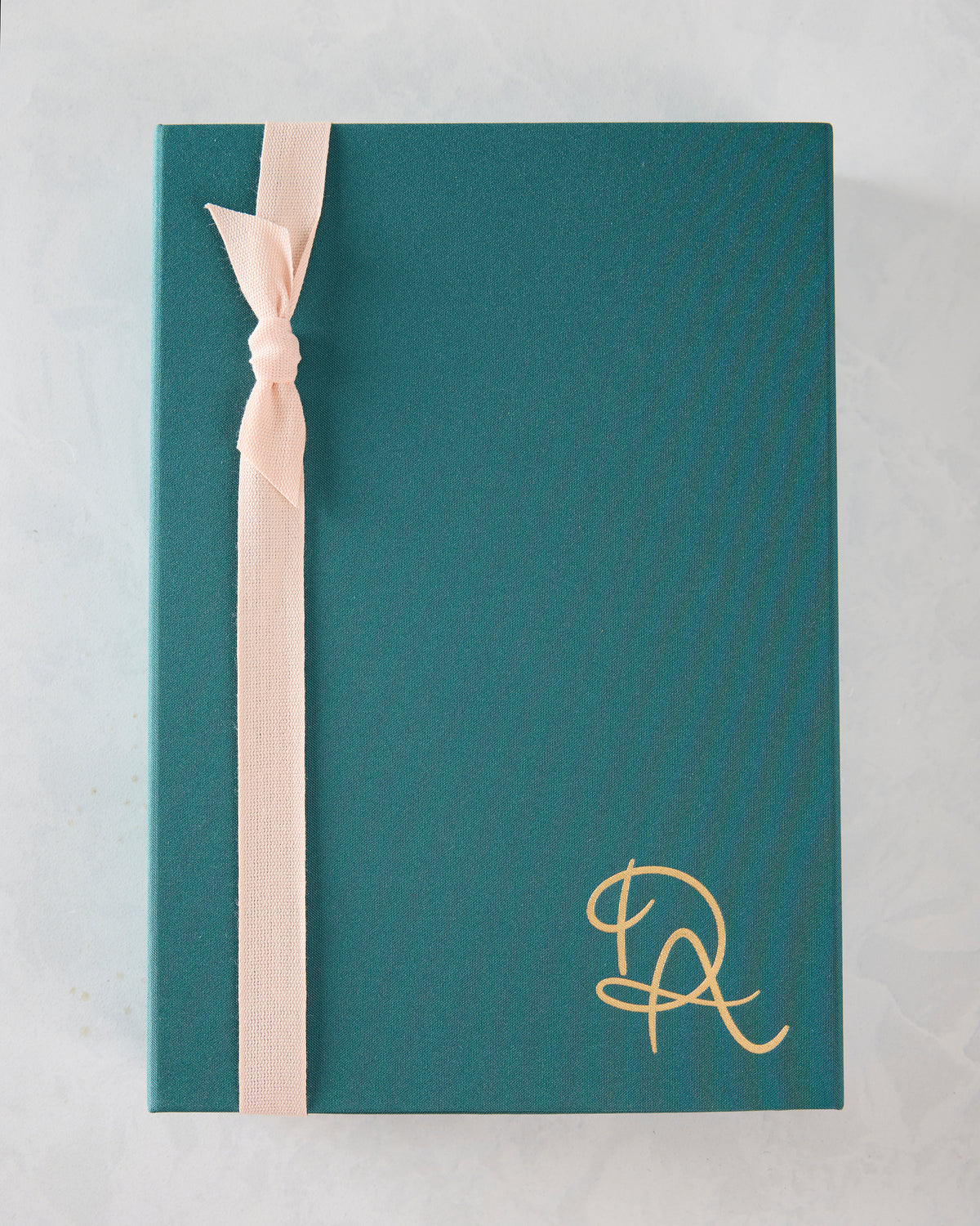Closed teal presentation box with a pale ribbon and gold monogram-style mark.
