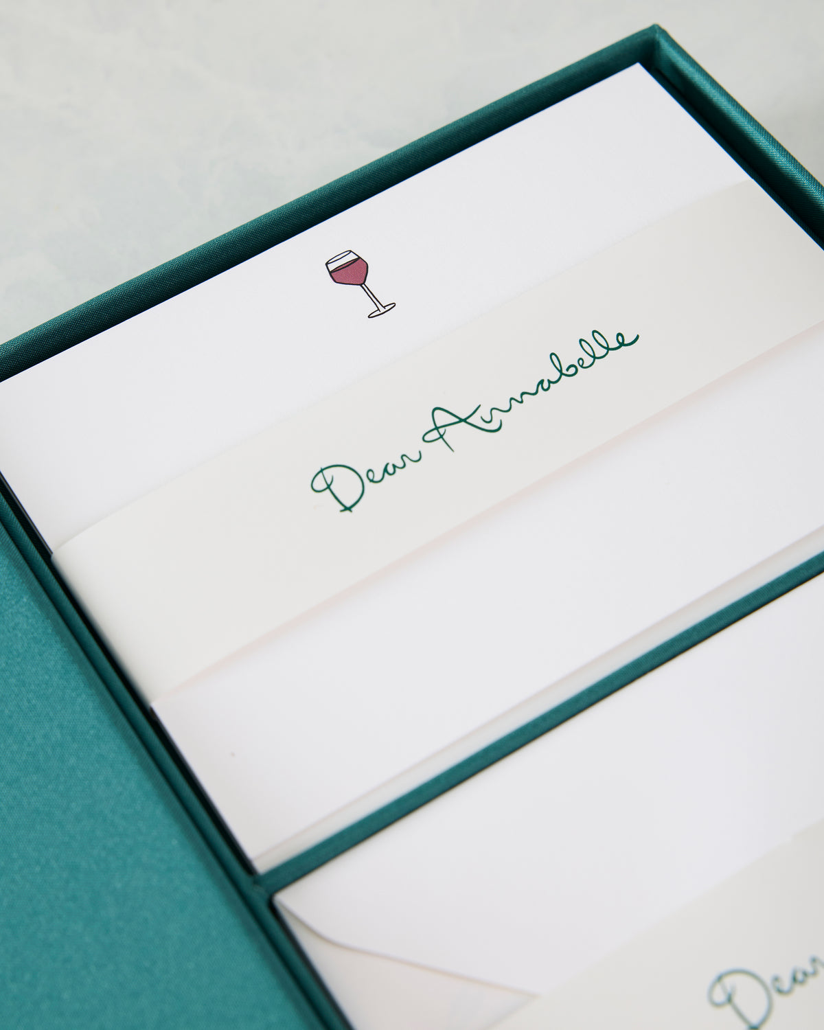 Close-up of a white notecard band reading “Dear Annabelle” beside a wine-glass icon inside the teal presentation box.