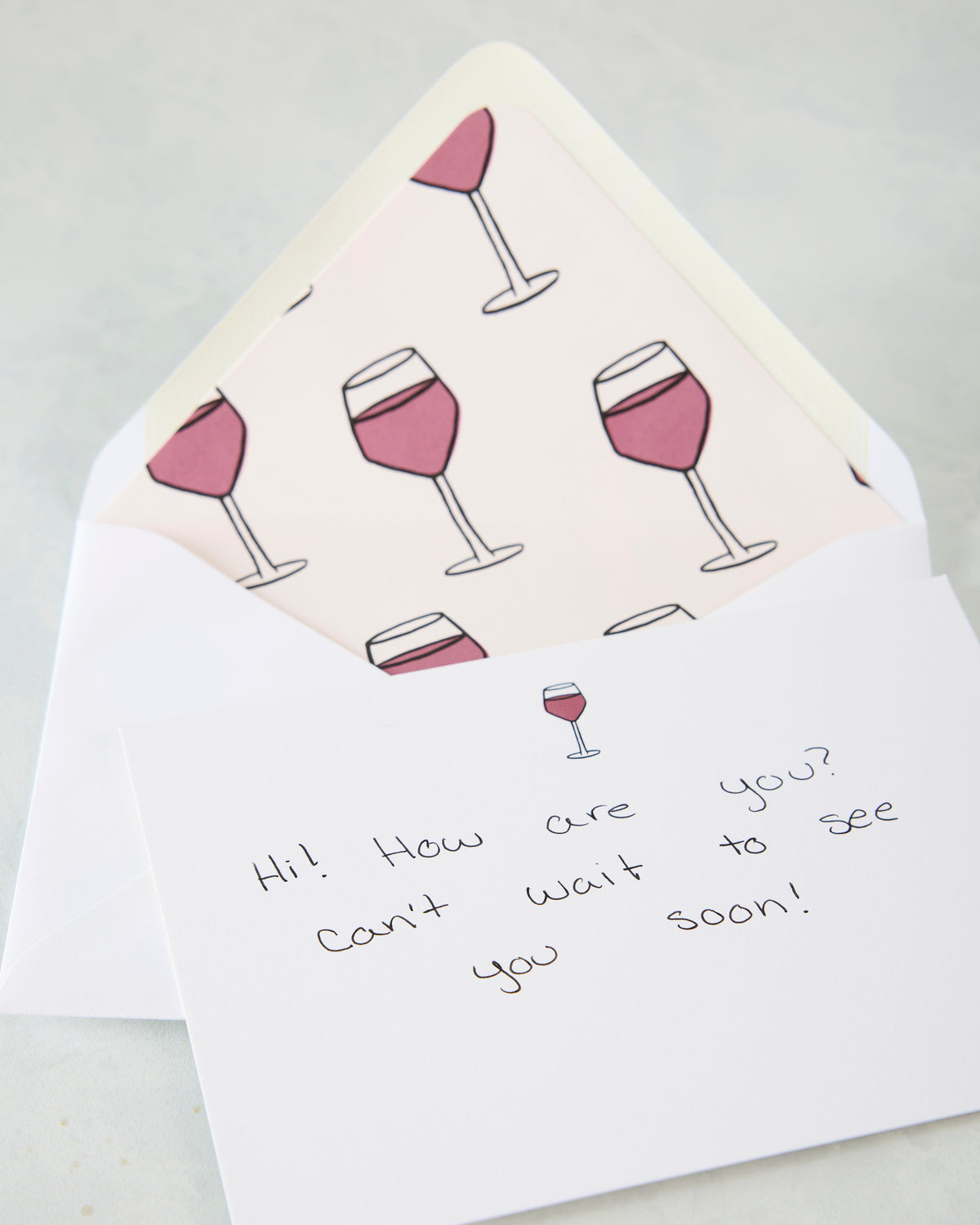 Open notecard and envelope showing a repeating wine-glass pattern; the handwritten note reads, “Hi! How are you? Can’t wait to see you soon!”