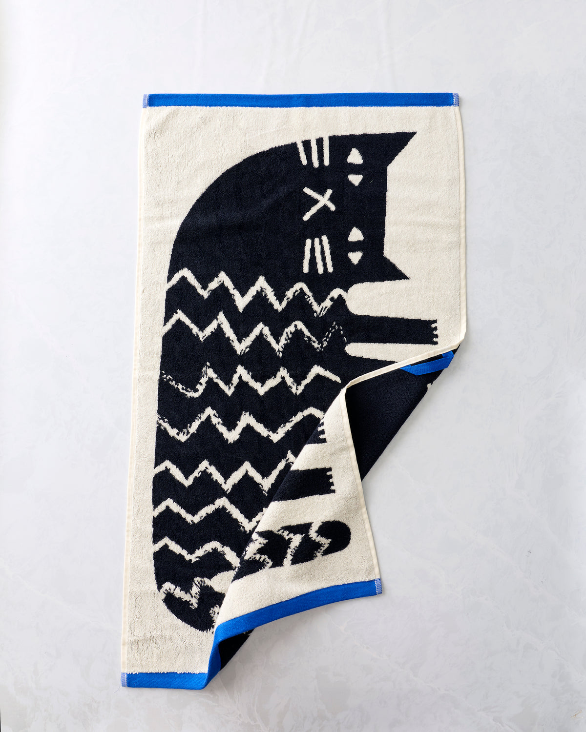 Black-and-cream hand towel with a stylized cat design, shown partially folded to reveal the reverse side and blue edging.