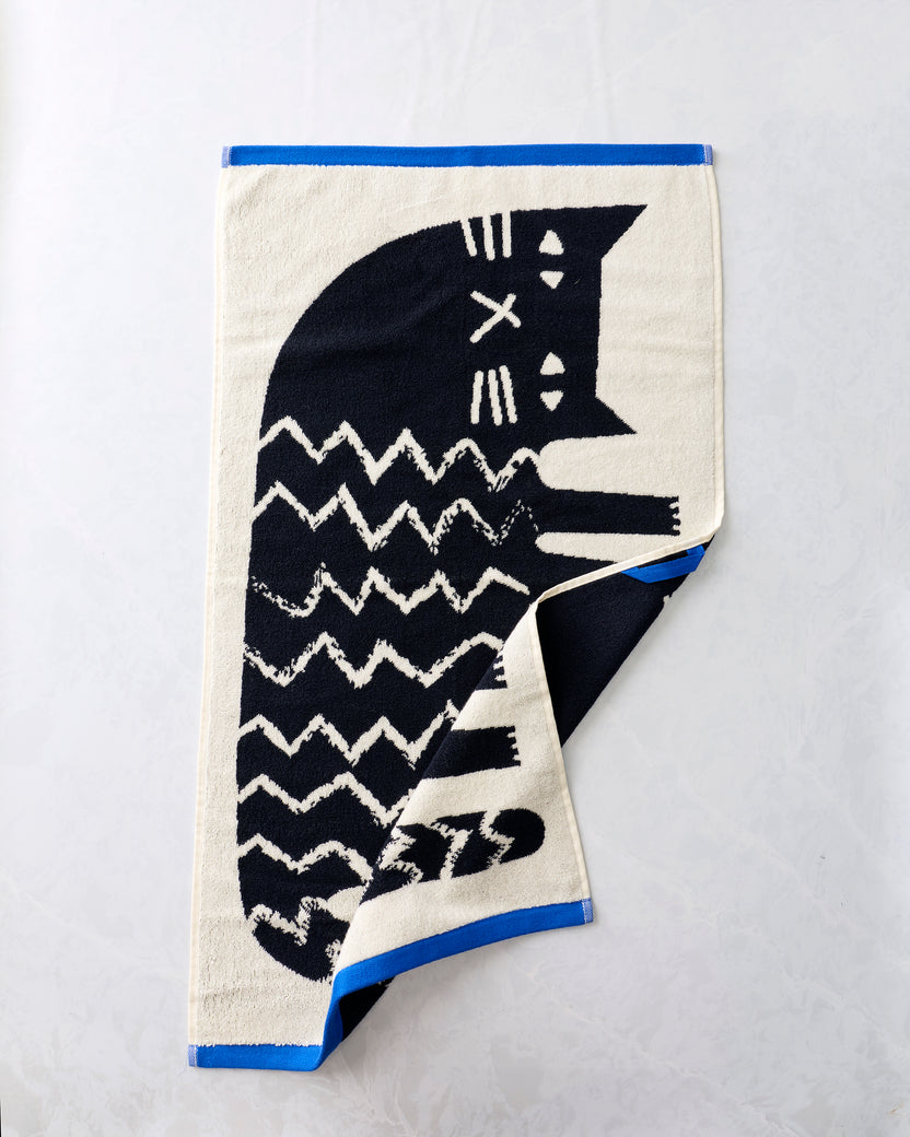 Black-and-cream hand towel with a stylized cat design, shown partially folded to reveal the reverse side and blue edging.