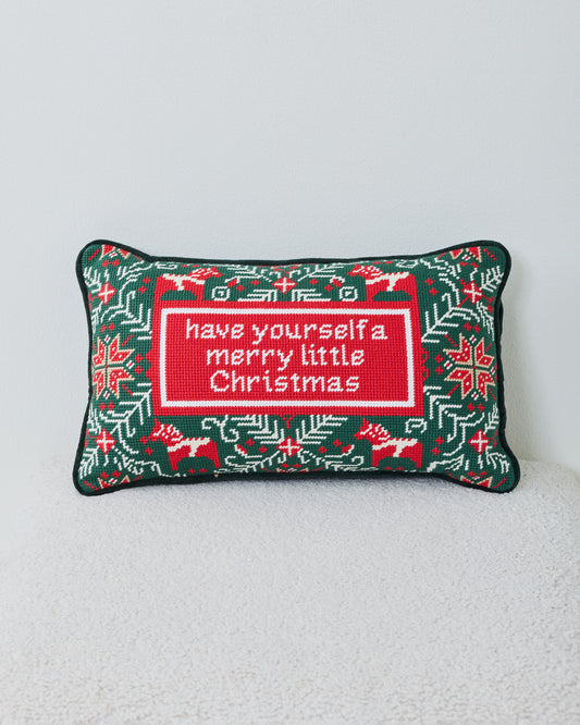 Front view of a green needlepoint Christmas pillow with reindeer, snowflakes, greenery, and a central holiday message.