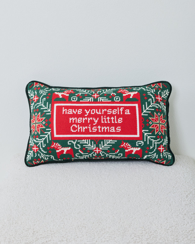 Front view of a green needlepoint Christmas pillow with reindeer, snowflakes, greenery, and a central holiday message.