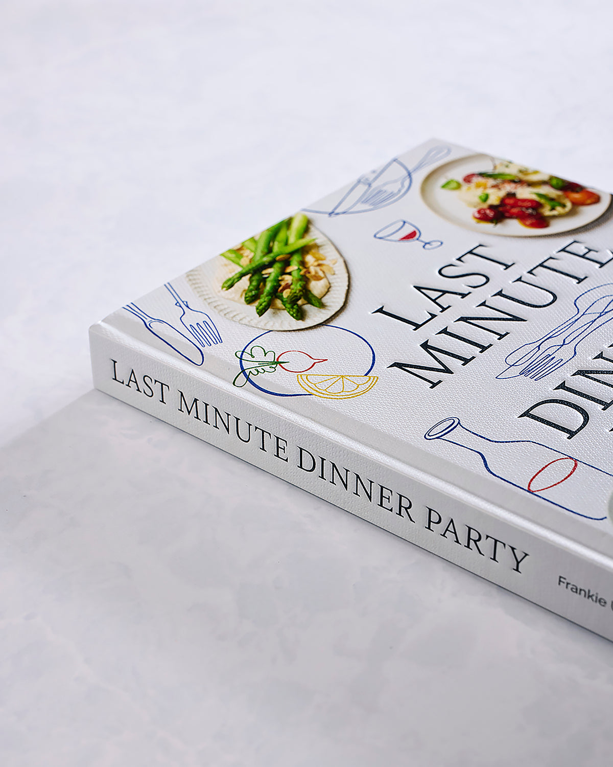 Angled view of the Last Minute Dinner Party cookbook, showing plated food photographs and colorful utensil and ingredient drawings.