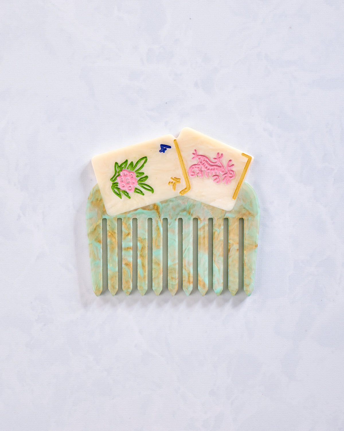 Green and cream decorative pocket comb with vertical teeth and painted floral and symbol details, shown on a light background.