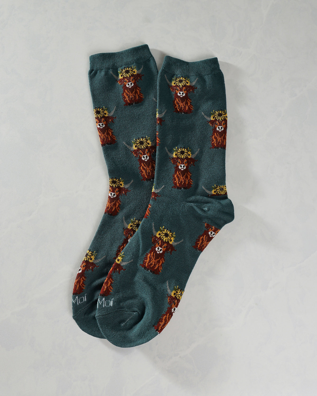 Teal Highland cow crew socks overlapping in a pair, with repeating flower-crowned cow illustrations.