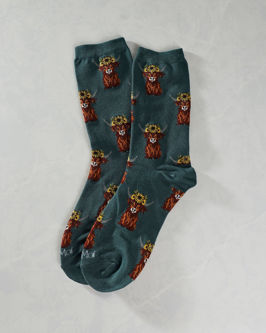 Teal Highland cow crew socks overlapping in a pair, with repeating flower-crowned cow illustrations.