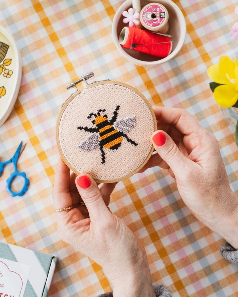 Hands hold a wooden hoop containing a black-and-yellow bumble bee cross-stitch design on peach fabric, with sewing supplies nearby.