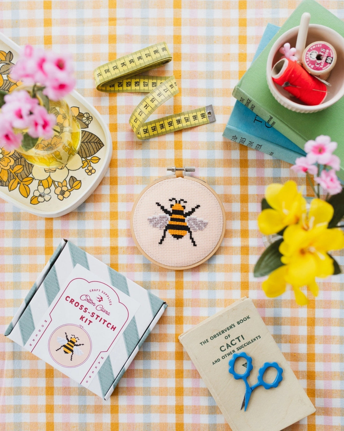 Bumble bee cross-stitch hoop displayed with the kit box, measuring tape, scissors, thread, books, and flowers on a gingham table.