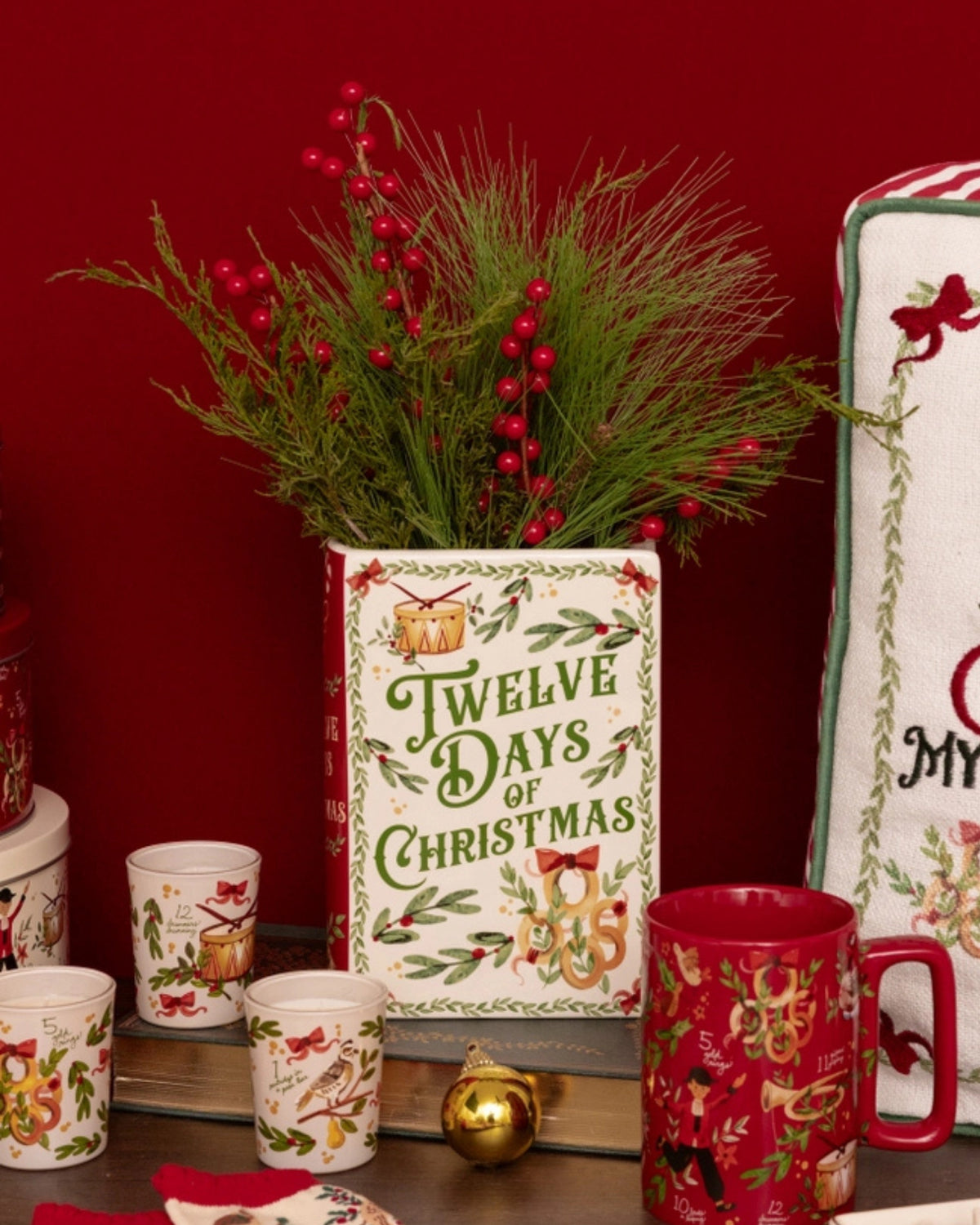 Twelve Days of Christmas book-style vase holding greenery with red berries, displayed among coordinating holiday cups and a mug.