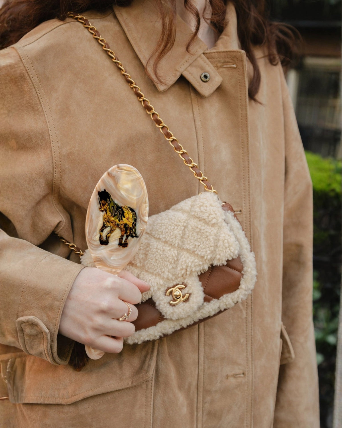 Hand-painted horse hair brush held beside a shearling-style handbag, shown with a person wearing a tan jacket.