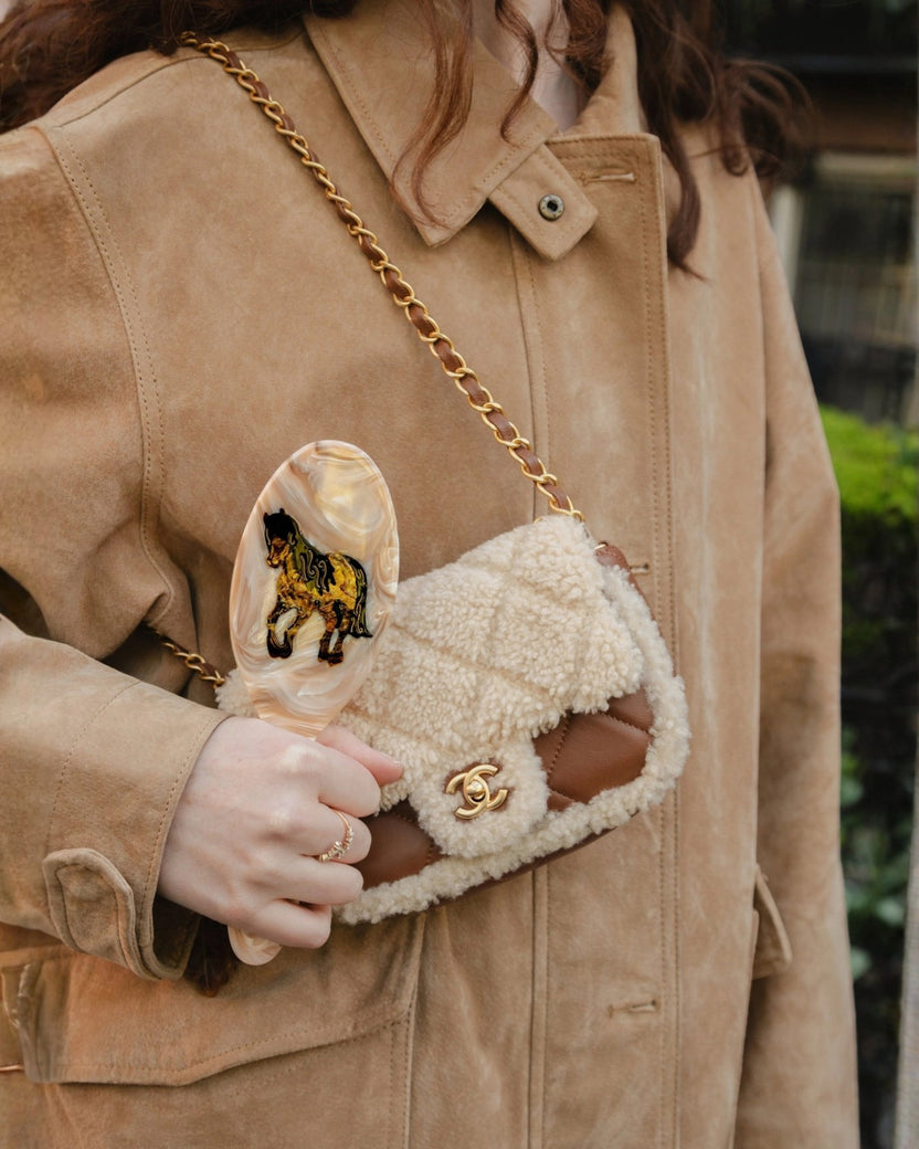 Hand-painted horse hair brush held beside a shearling-style handbag, shown with a person wearing a tan jacket.