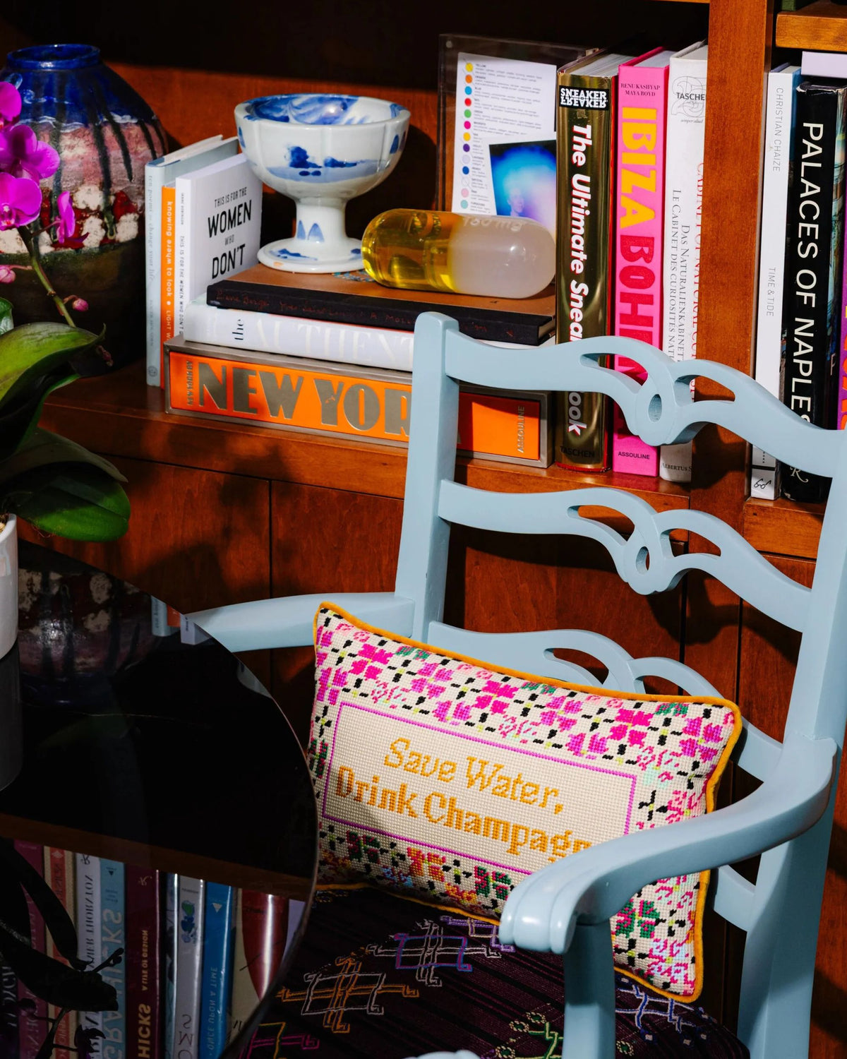 Needlepoint “Save Water, Drink Champagne” pillow displayed on a light blue chair beside a bookcase.
