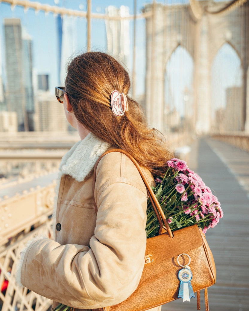 Person wearing the coffee-cup-shaped hair clip in a ponytail on a bridge, with a brown handbag and flowers.