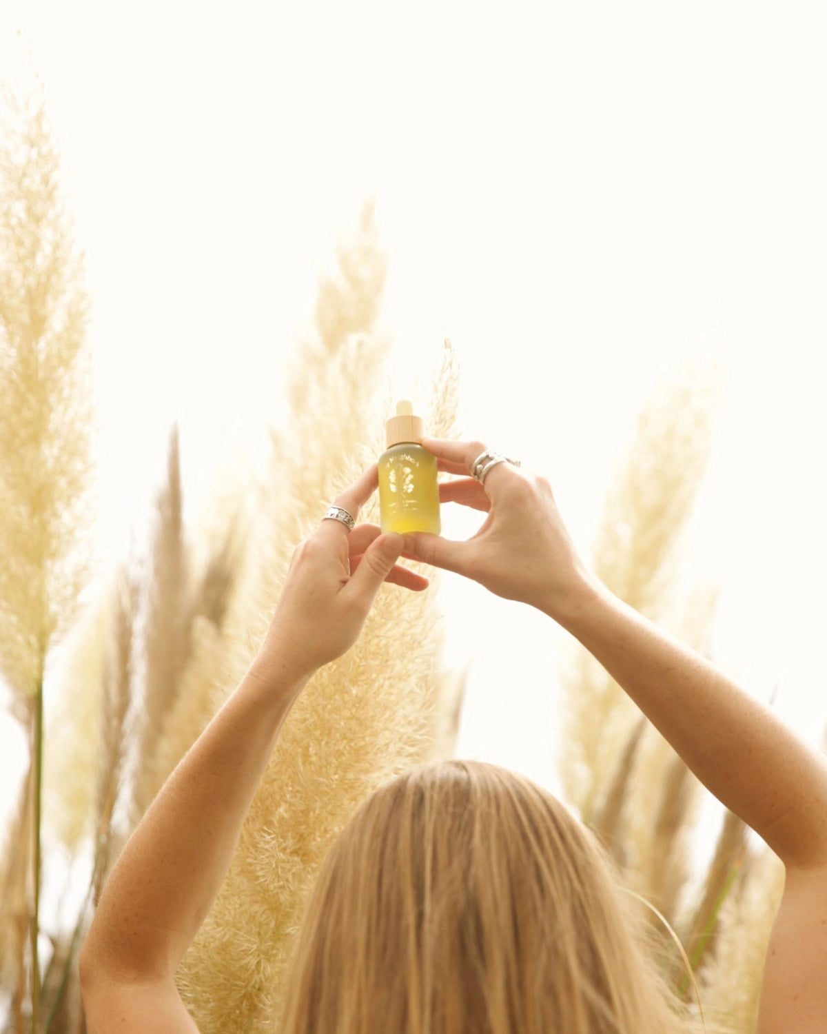 Two hands hold the Rosemary & Juniper Hair Oil dropper bottle against a bright field of tall grass.