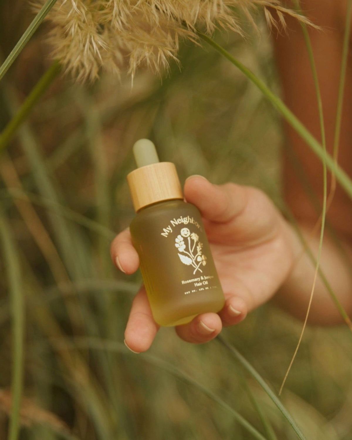 Close view of a hand holding the My Neighbor's Rosemary & Juniper Hair Oil dropper bottle among tall grass.