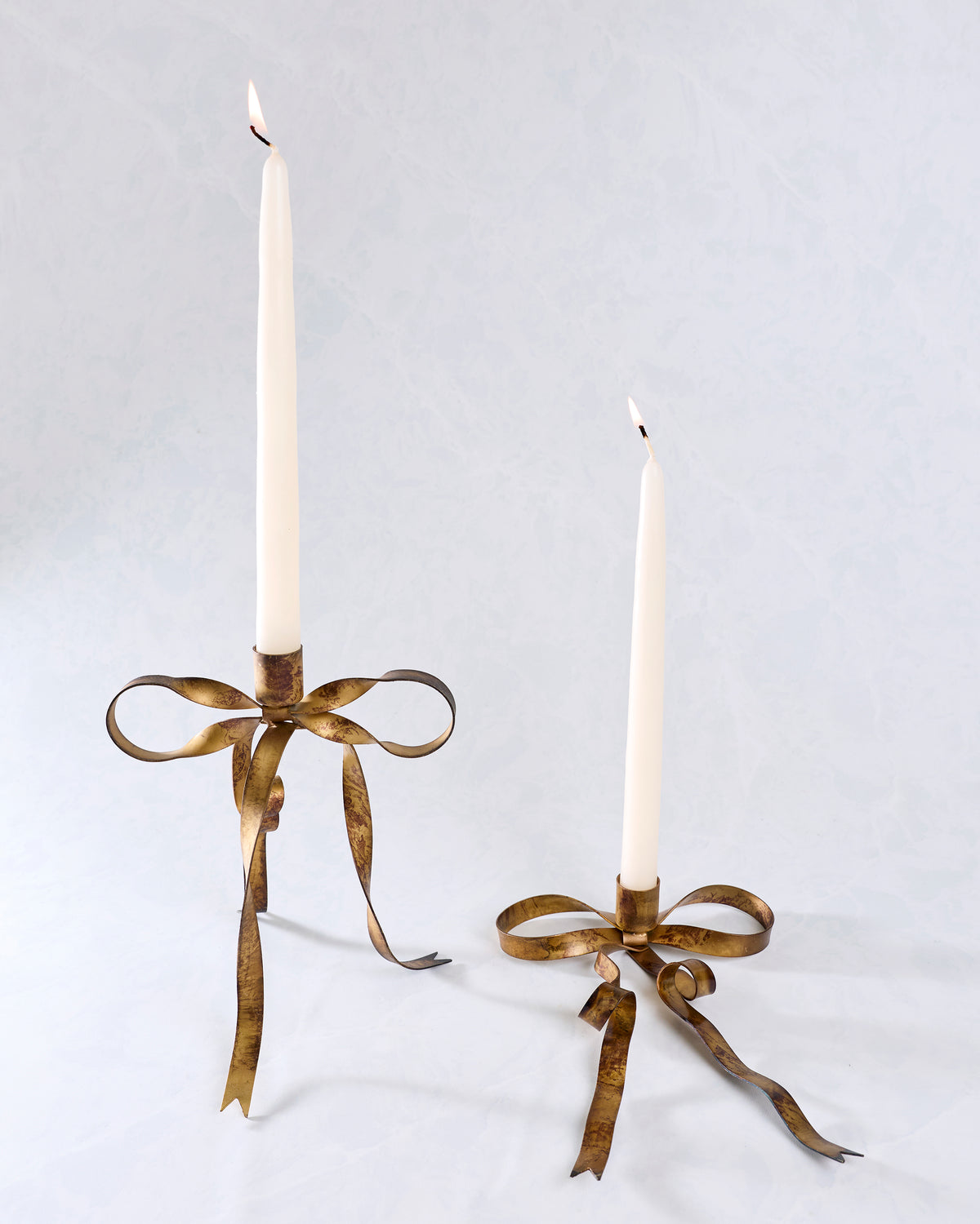Two brass bow-shaped taper candle holders in different sizes, each holding a lit white taper candle.