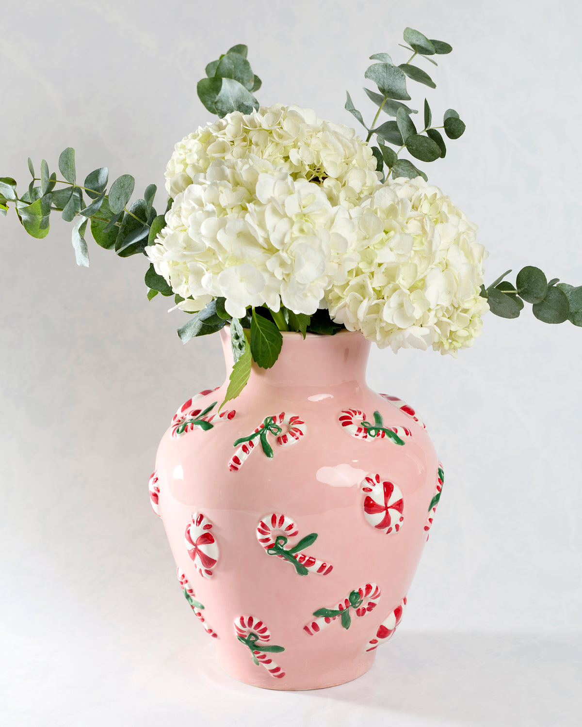 Pink ceramic vase with raised red-and-green peppermint details, holding white flowers and eucalyptus greenery.