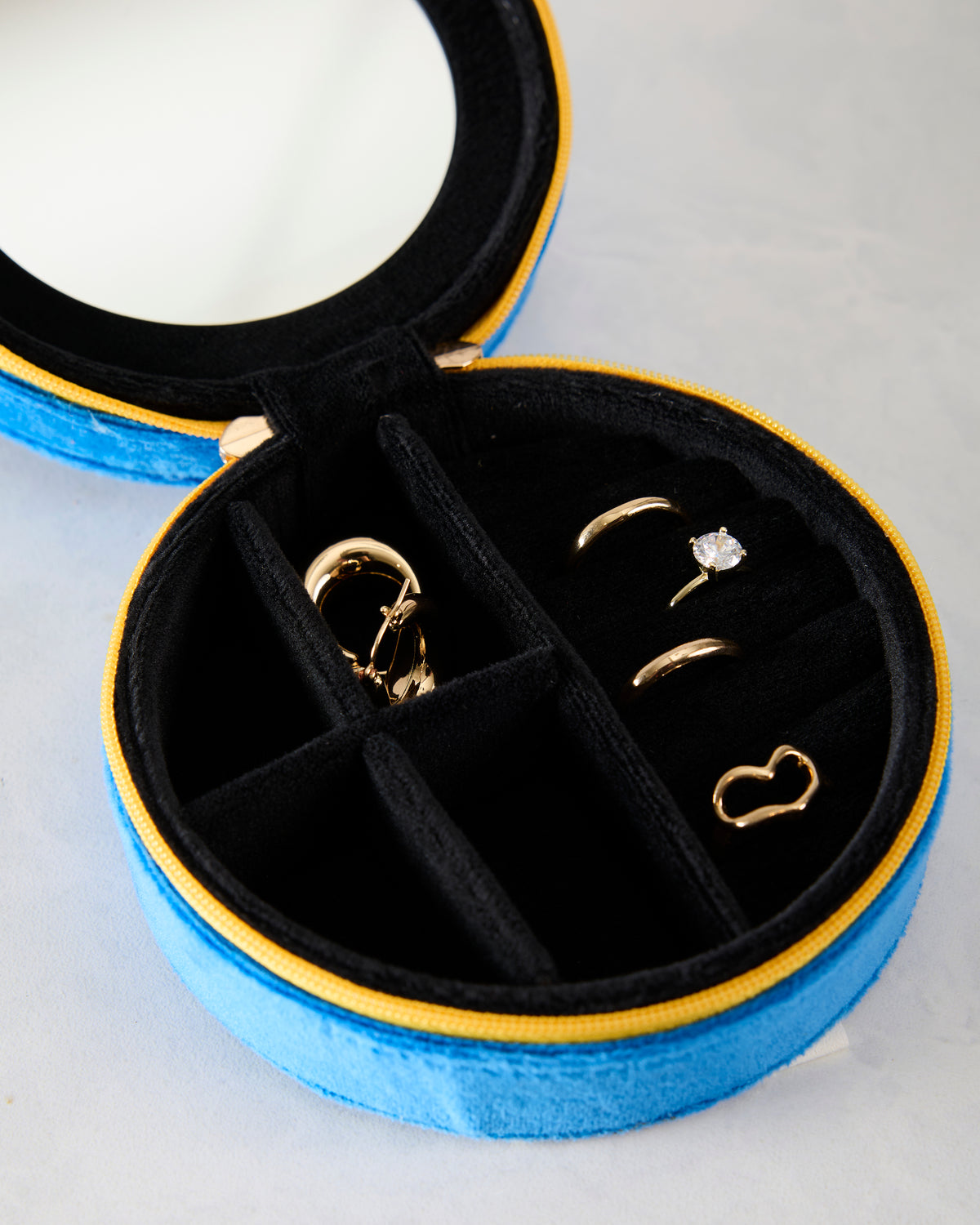 Open blue velvet jewelry zip case with black compartments, a round mirror, and several gold-tone jewelry pieces.