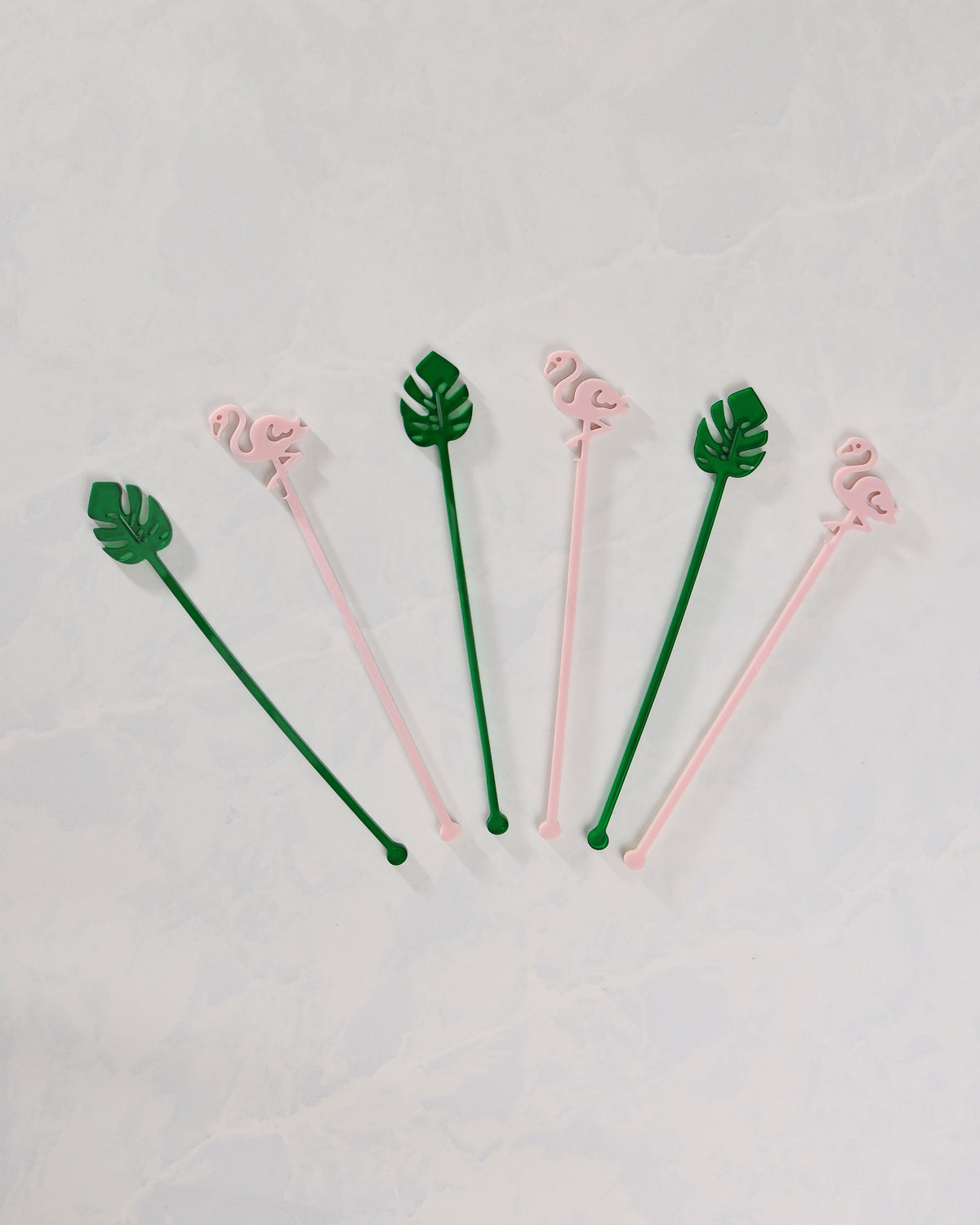 Six pink flamingo and green palm-leaf stir sticks arranged separately on a white surface.