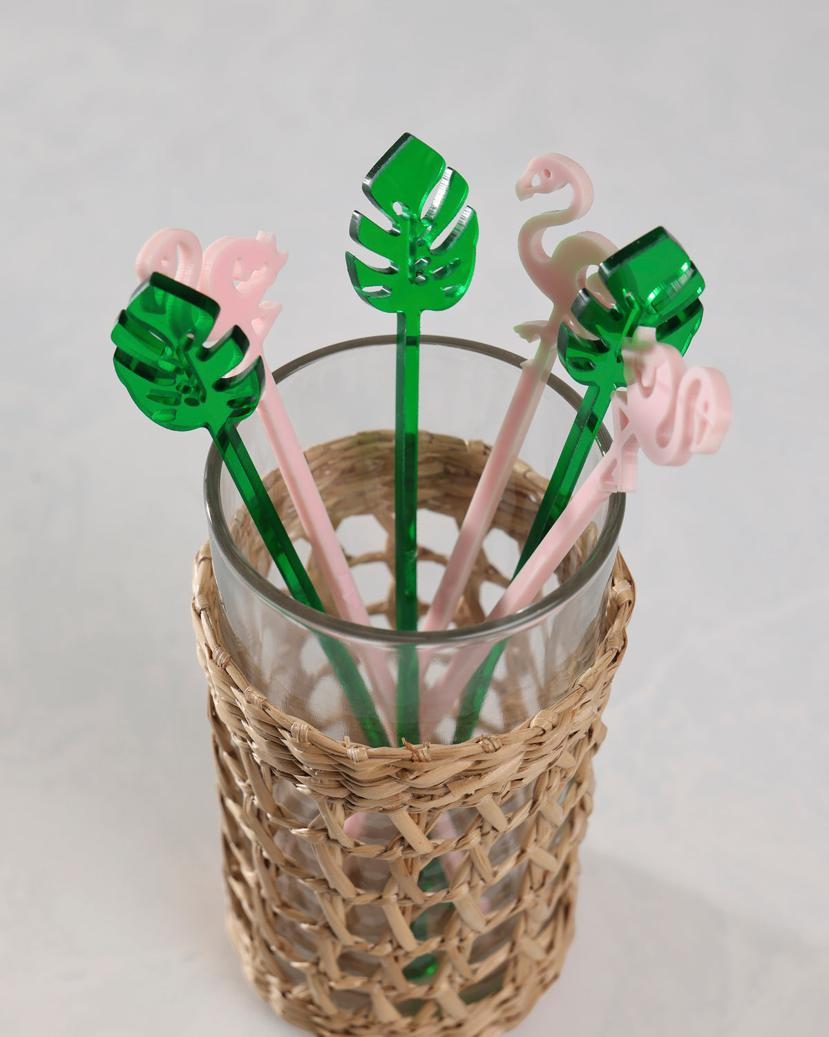 Pink flamingo and green palm-leaf stir sticks displayed in a glass inside a woven holder.