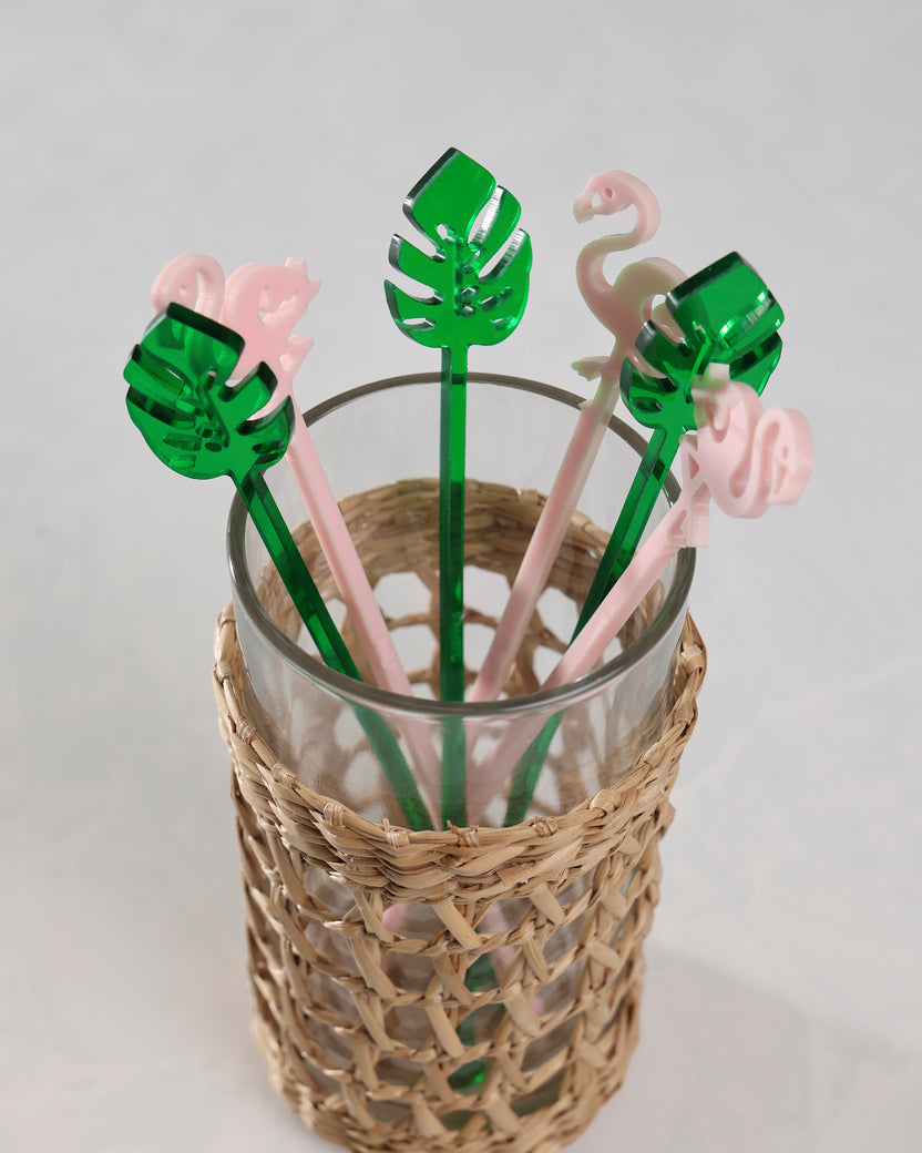 Pink flamingo and green palm-leaf stir sticks displayed in a glass inside a woven holder.