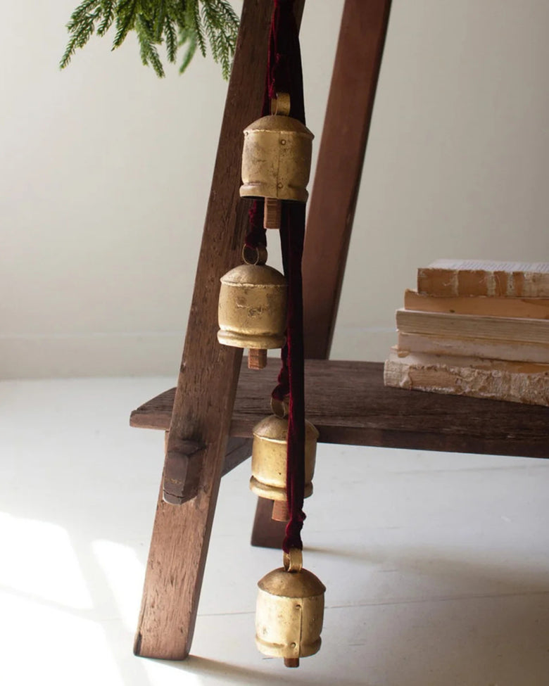 Four gold jingle bells hanging vertically from a maroon ribbon beside a wooden shelf.