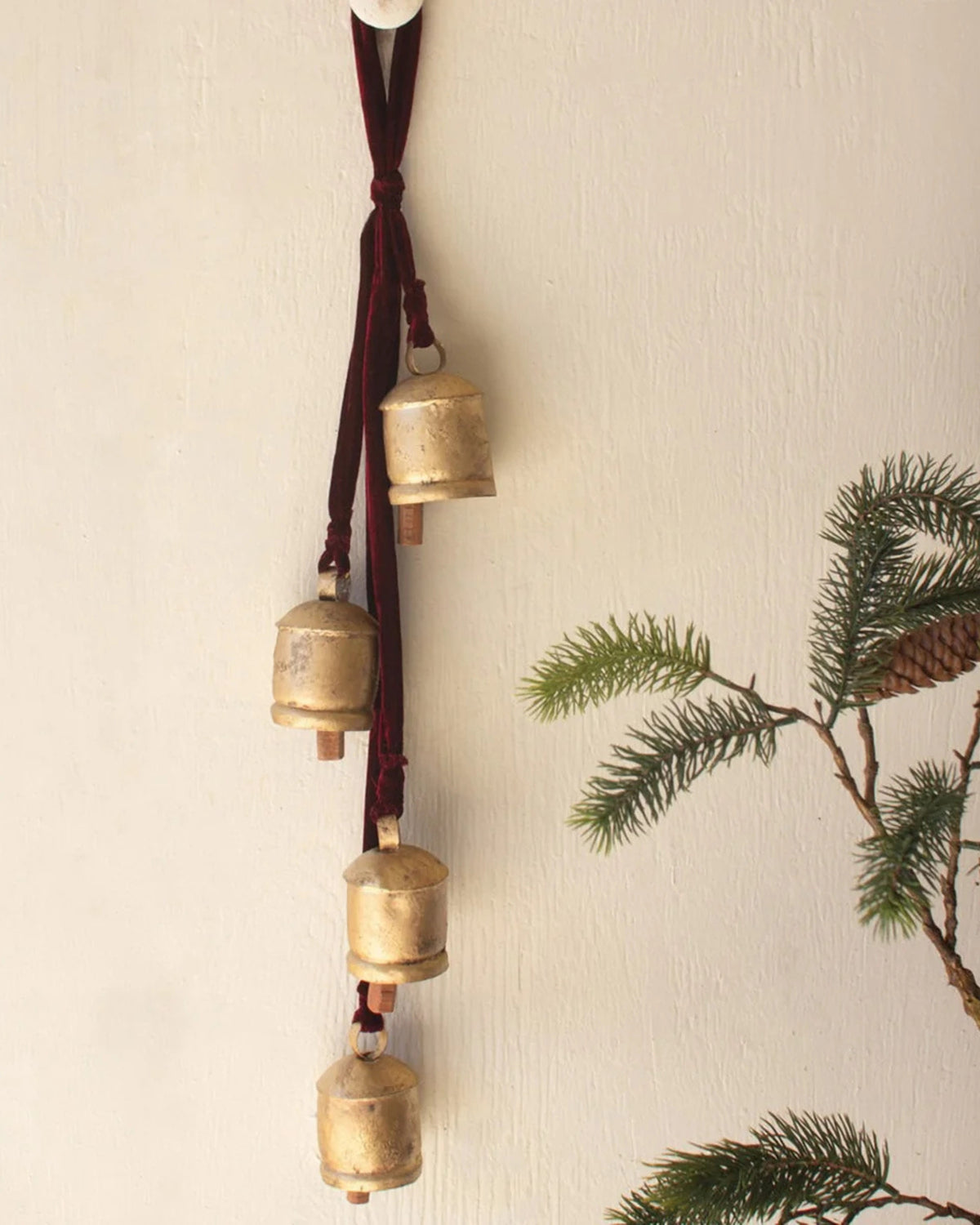 Gold jingle bells hanging vertically from a maroon ribbon on a wall beside evergreen branches.
