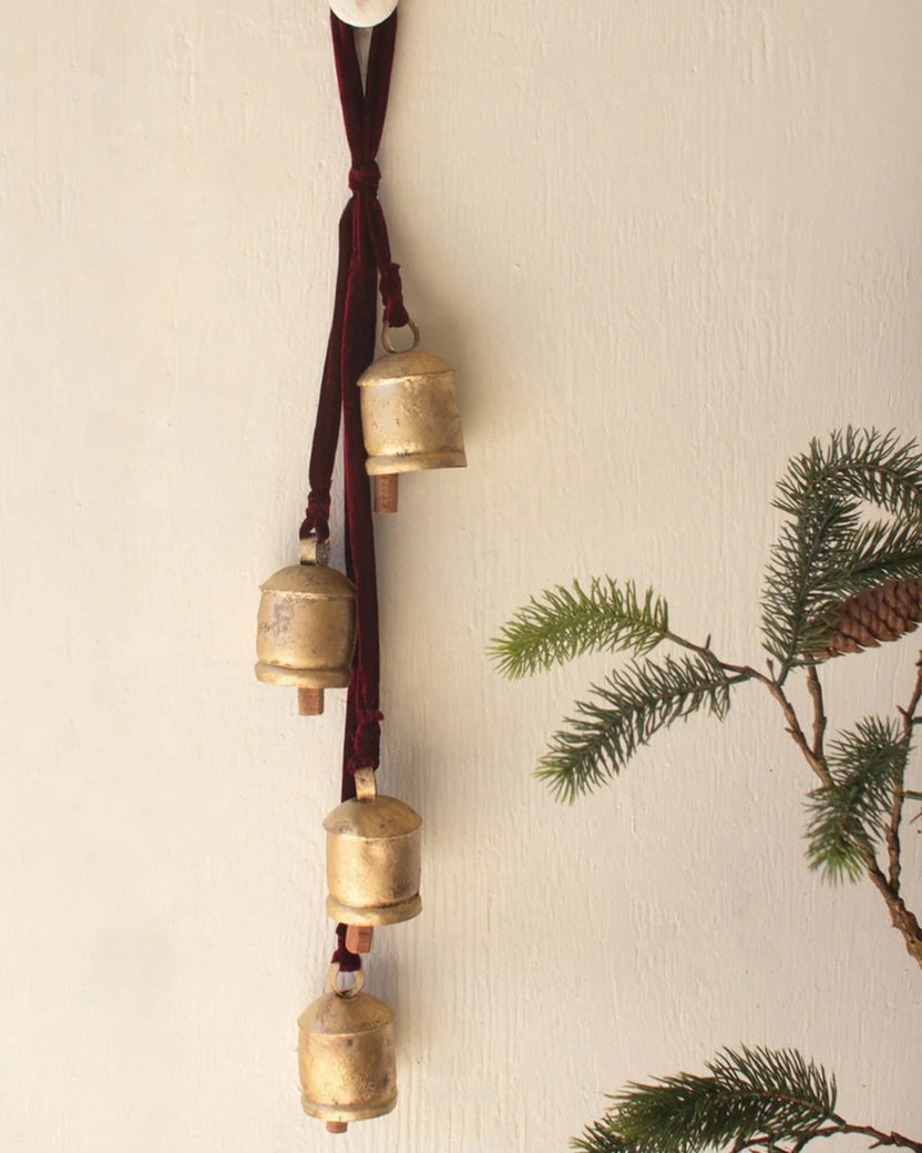Gold jingle bells hanging vertically from a maroon ribbon on a wall beside evergreen branches.