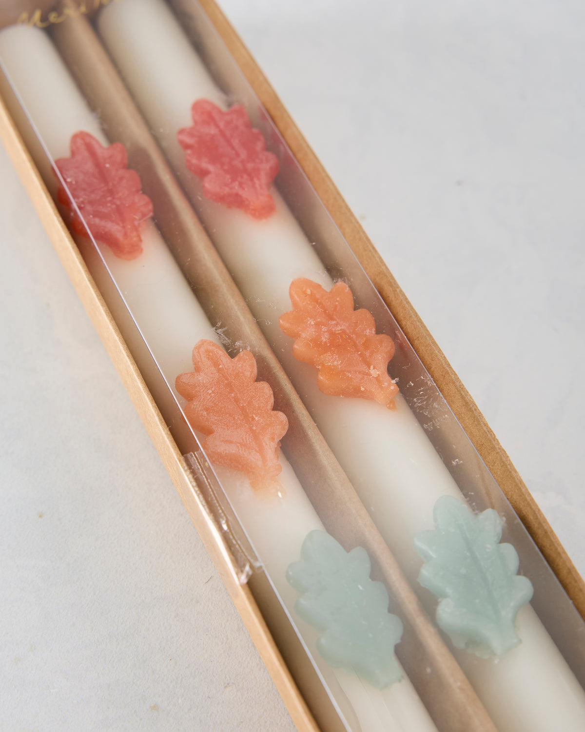 Angled close-up of two white candles in a box, decorated with pink, orange, and pale green leaf-shaped accents.