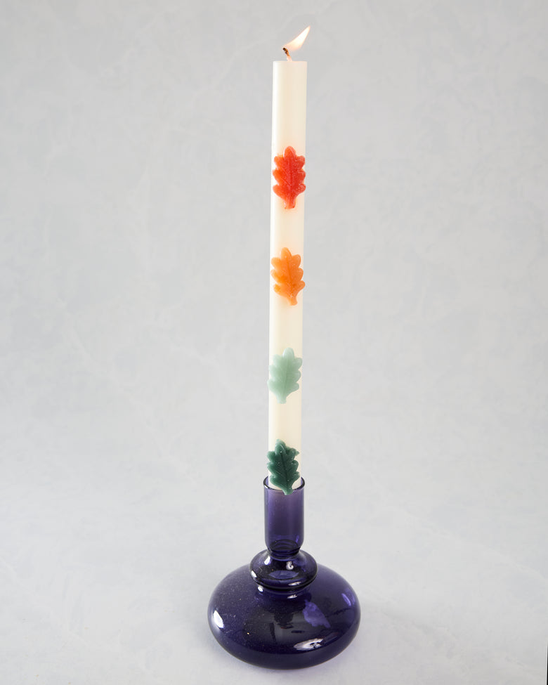 Full view of a lit white leaf-decorated candle in a dark purple glass holder, with red-orange, orange, pale green, and dark green leaf accents.