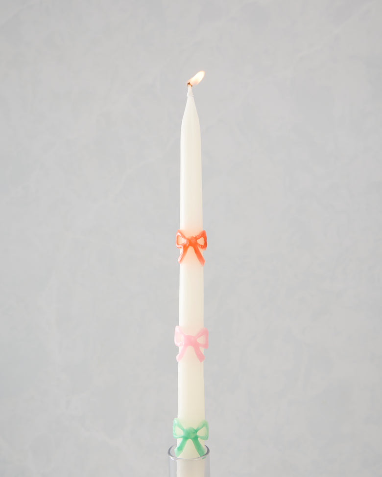 Lit white taper candle with orange, pink, and green bow decorations in a candle holder.