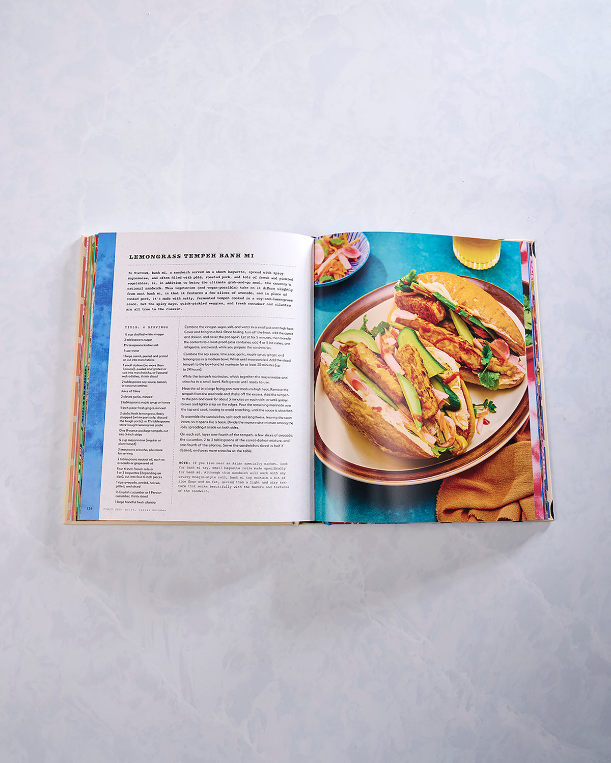 Open cookbook showing the recipe “Lemongrass Tempeh Banh Mi” beside a photograph of two filled sandwiches.