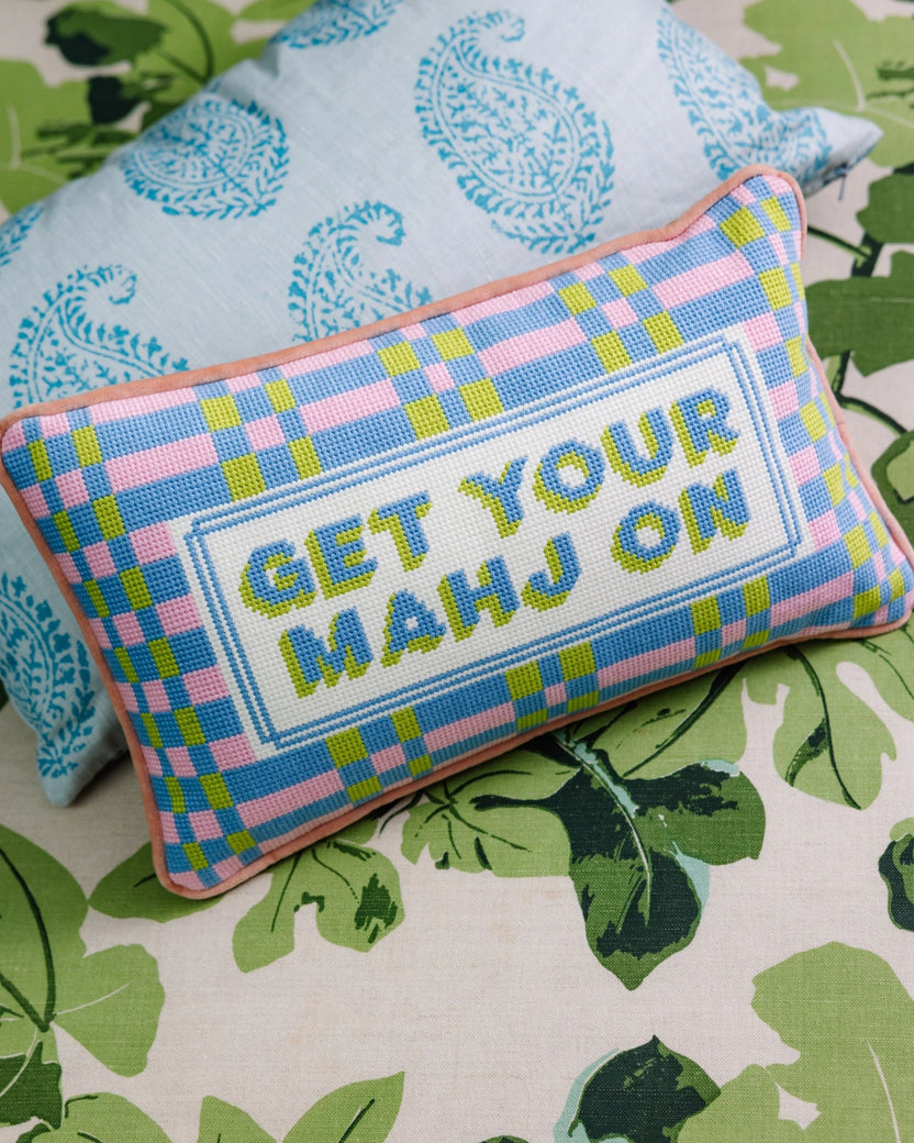 Needlepoint pillow reading “GET YOUR MAHJ ON,” angled on a bed with botanical-patterned bedding.
