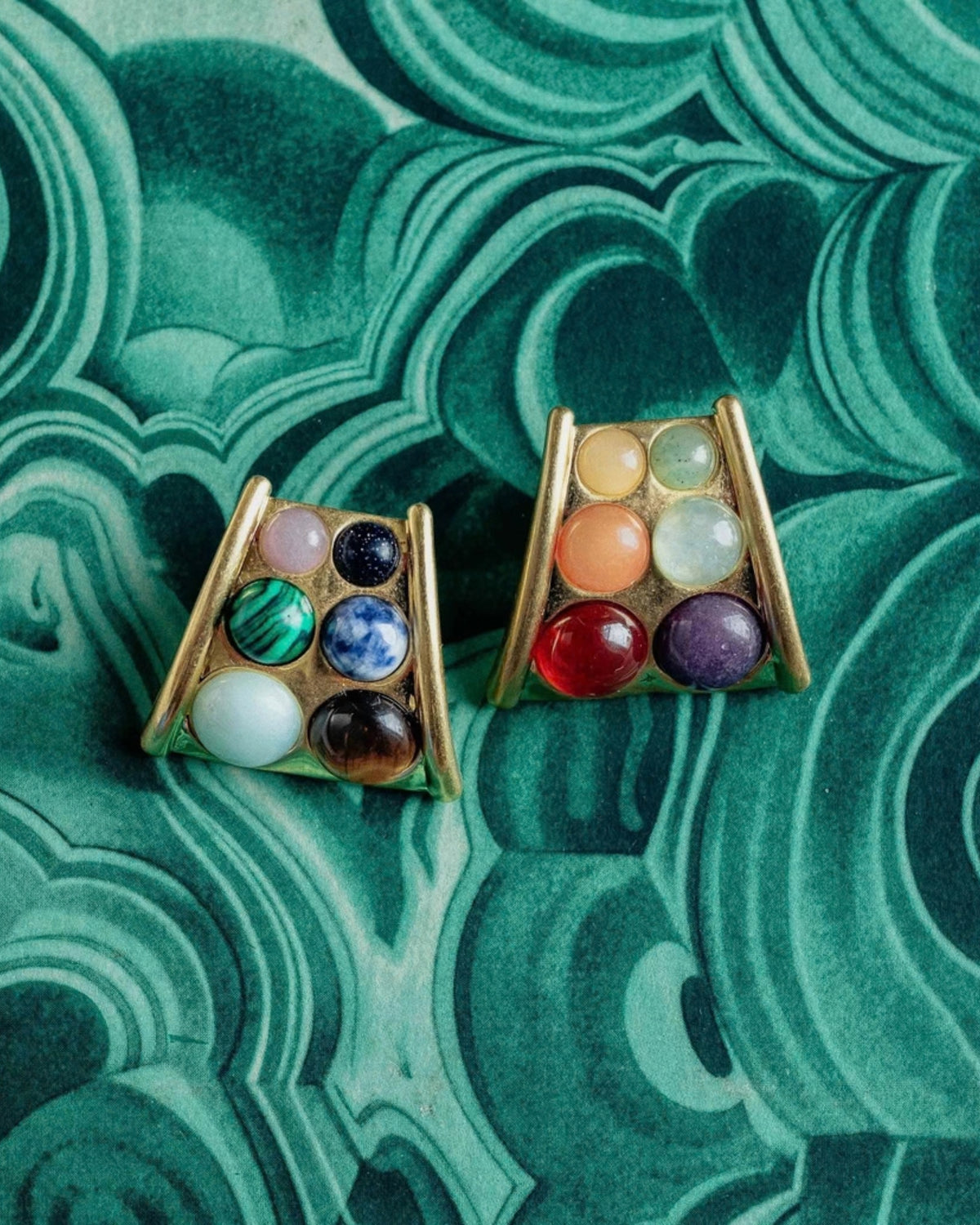 Pair of triangular gold-tone statement earrings with six multicolored round stones in each earring, shown on a green patterned background.