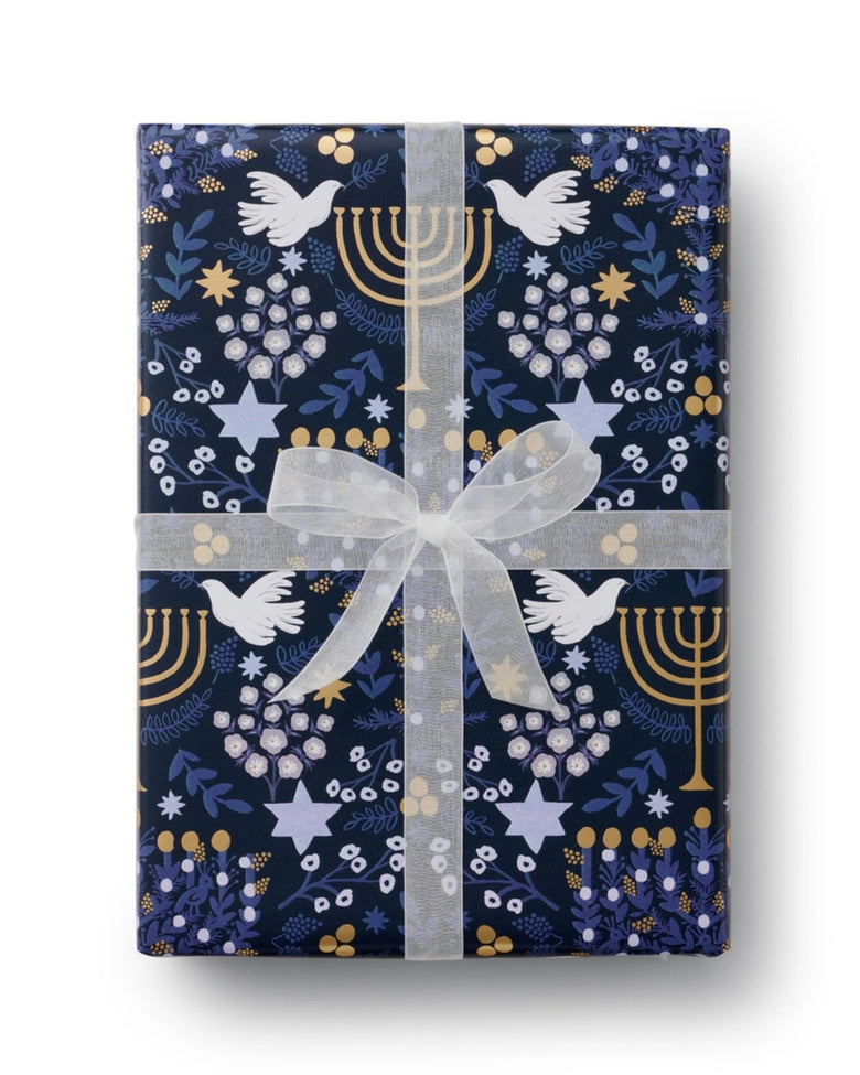 Navy Laurel Menorah wrapping paper wrapped as a gift with a translucent white ribbon and bow; the pattern includes doves, menorahs, stars, leaves, and berries.
